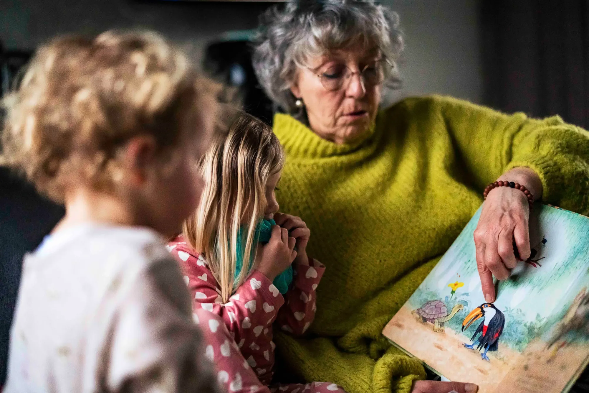 Vrouw en kinderen kijken samen in een prentenboek met kleurrijke dieren, terwijl ze binnen zitten, mogelijk in een bibliotheek of klaslokaal.