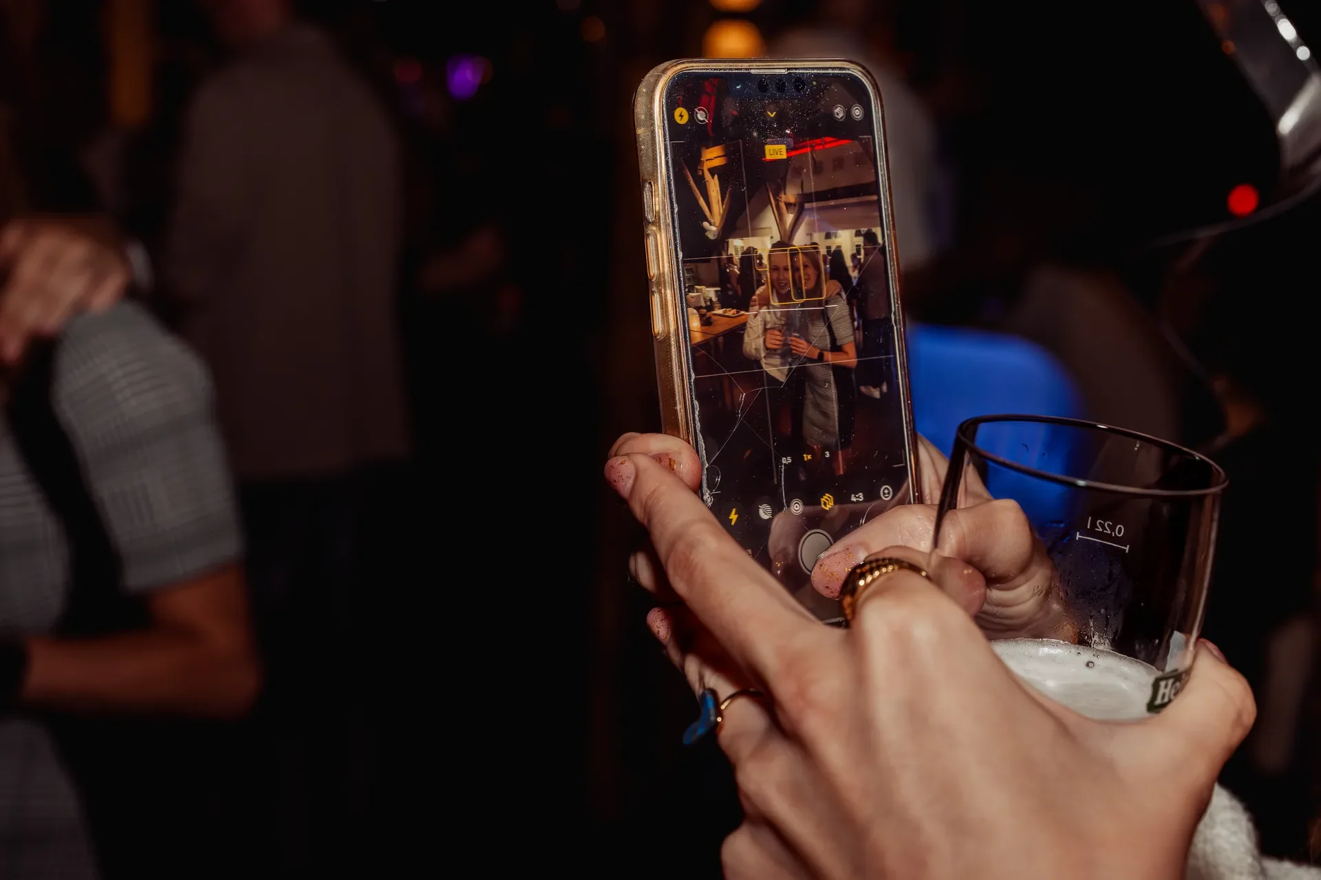 Persoon houdt een smartphone vast die een foto maakt van een vrouw met een drankje in een drukke, donkere bar of club.