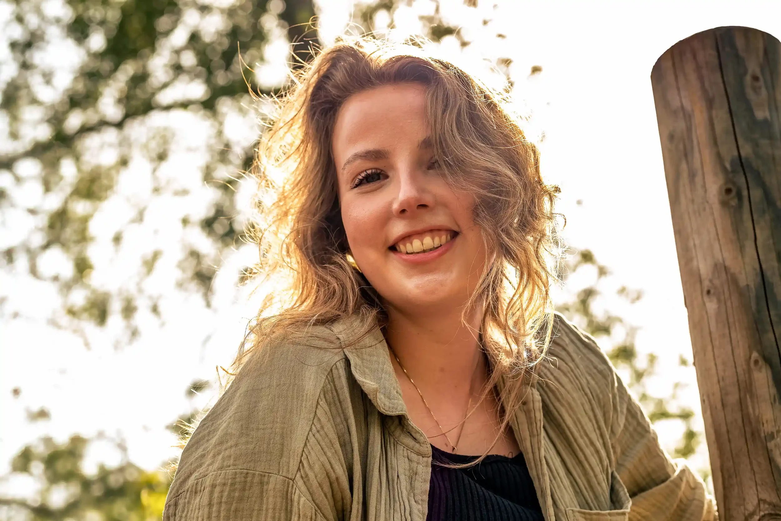 Een glimlachende jonge vrouw met golvend haar, zittend naast een houten paal, buitenshuis met bomen op de achtergrond en de zon die erachter schijnt.
