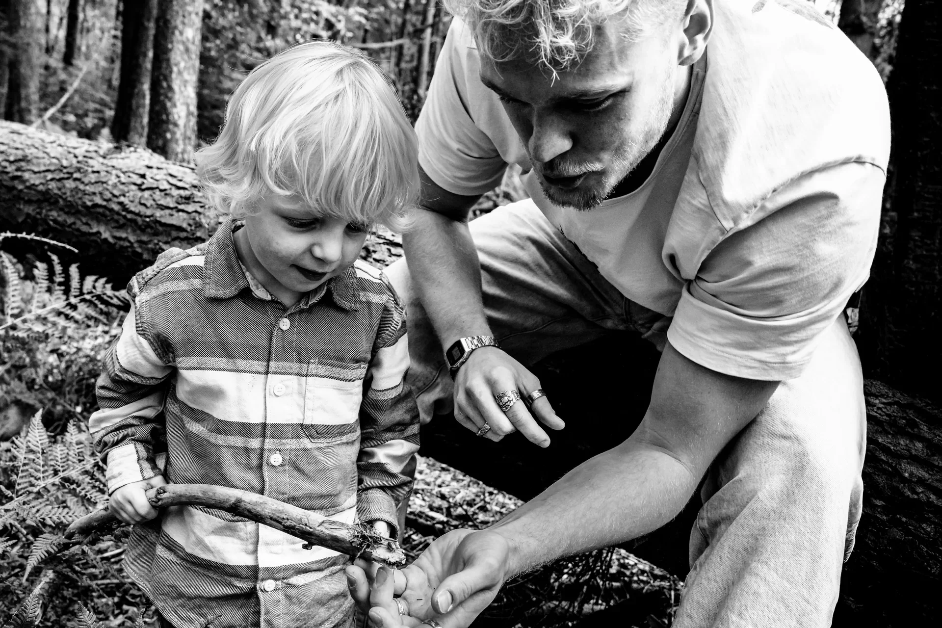 Een man en een jongen die samen iets onderzoeken in een bos, de man helpt de jongen met een stuk hout of tak.