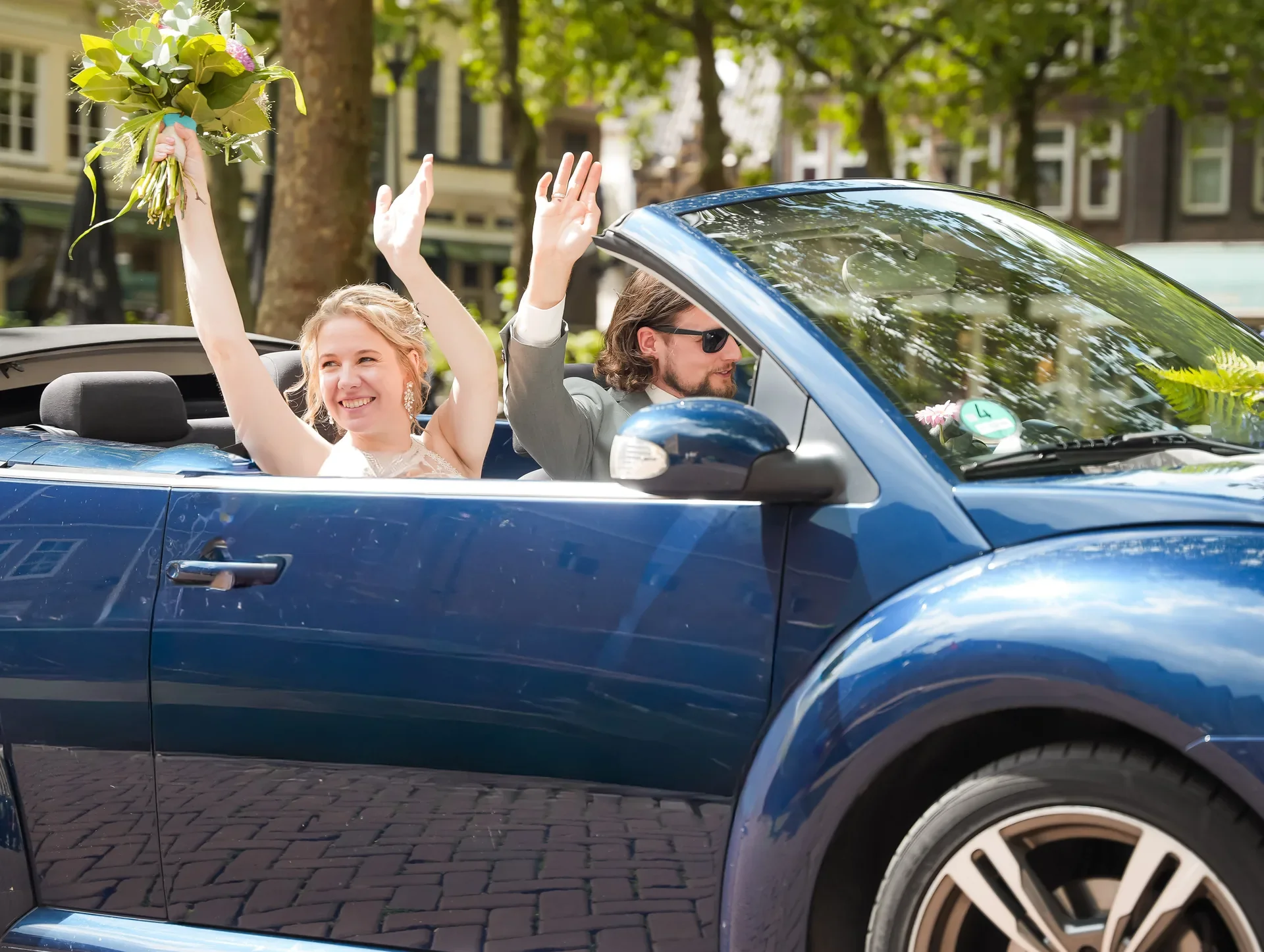 Een bruid en bruidegom rijden in een blauwe cabriolet, de bruid zwaait met beide handen en houdt een bloemenboeket omhoog, de bruidegom draagt zonnebril en groet bovendien.