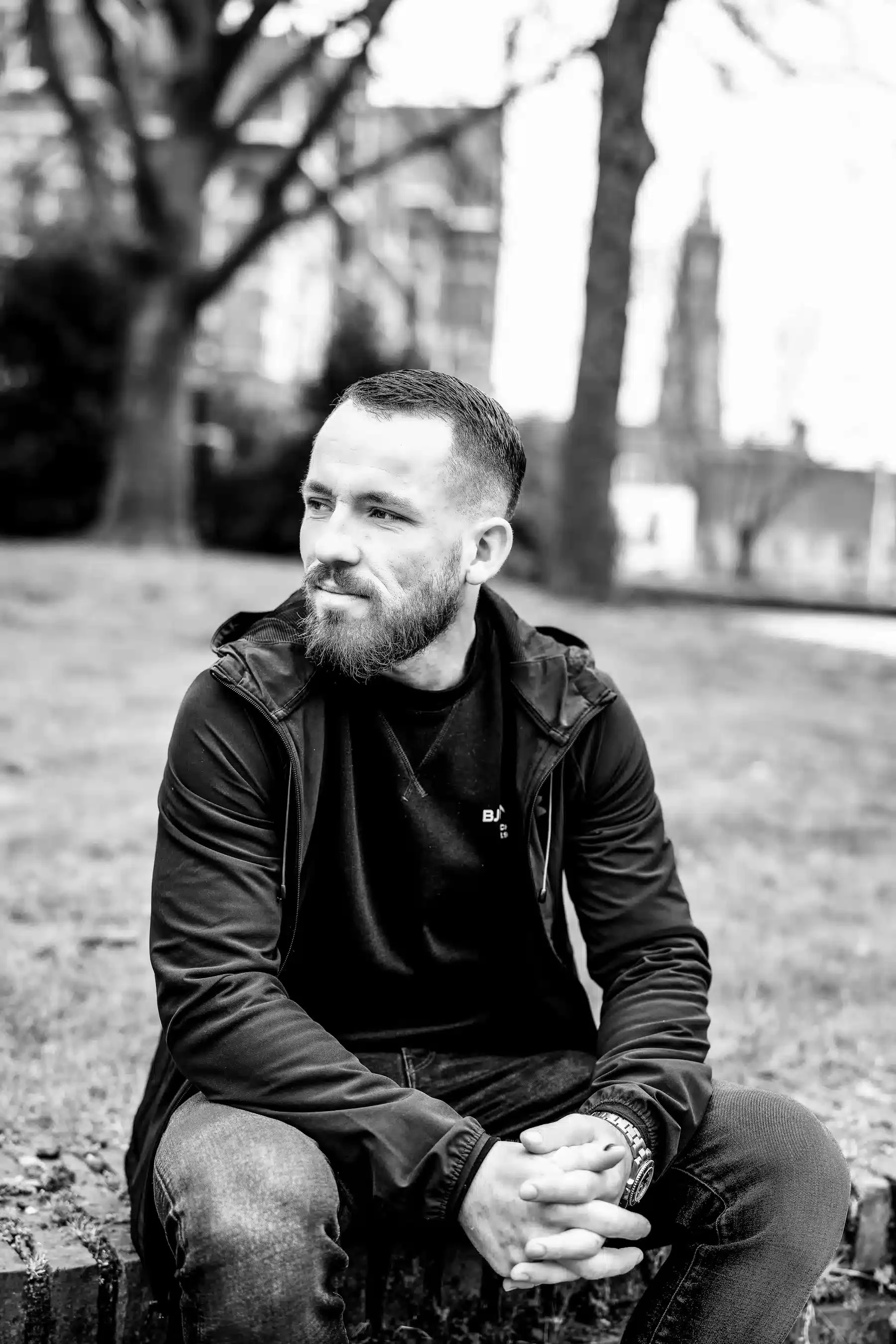 Zwart-wit foto van een man met een baard, zittend op een bankje in een park met bomen op de achtergrond, kijkend naar links.