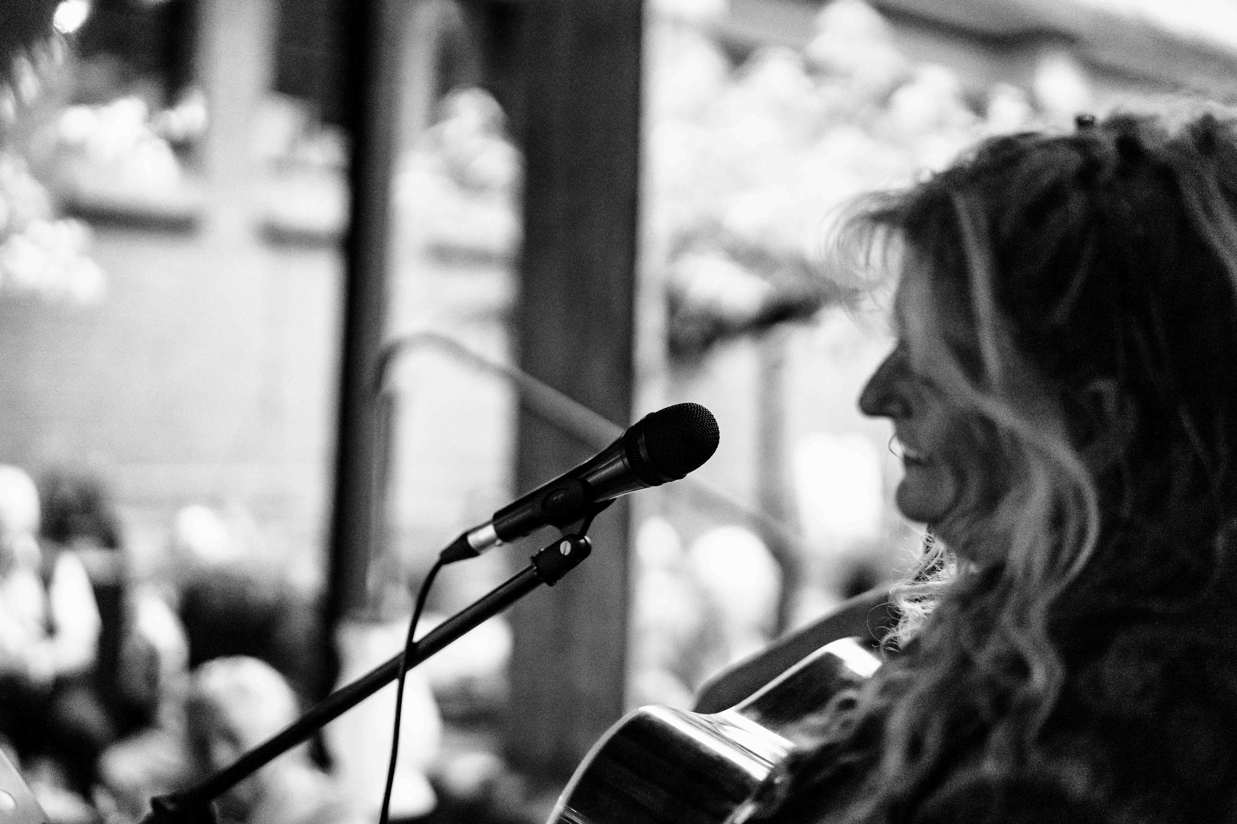 Een vrouw met golvend haar speelt gitaar en zingt in een microfoon, op de achtergrond mensen die luisteren, alles in zwart-wit.