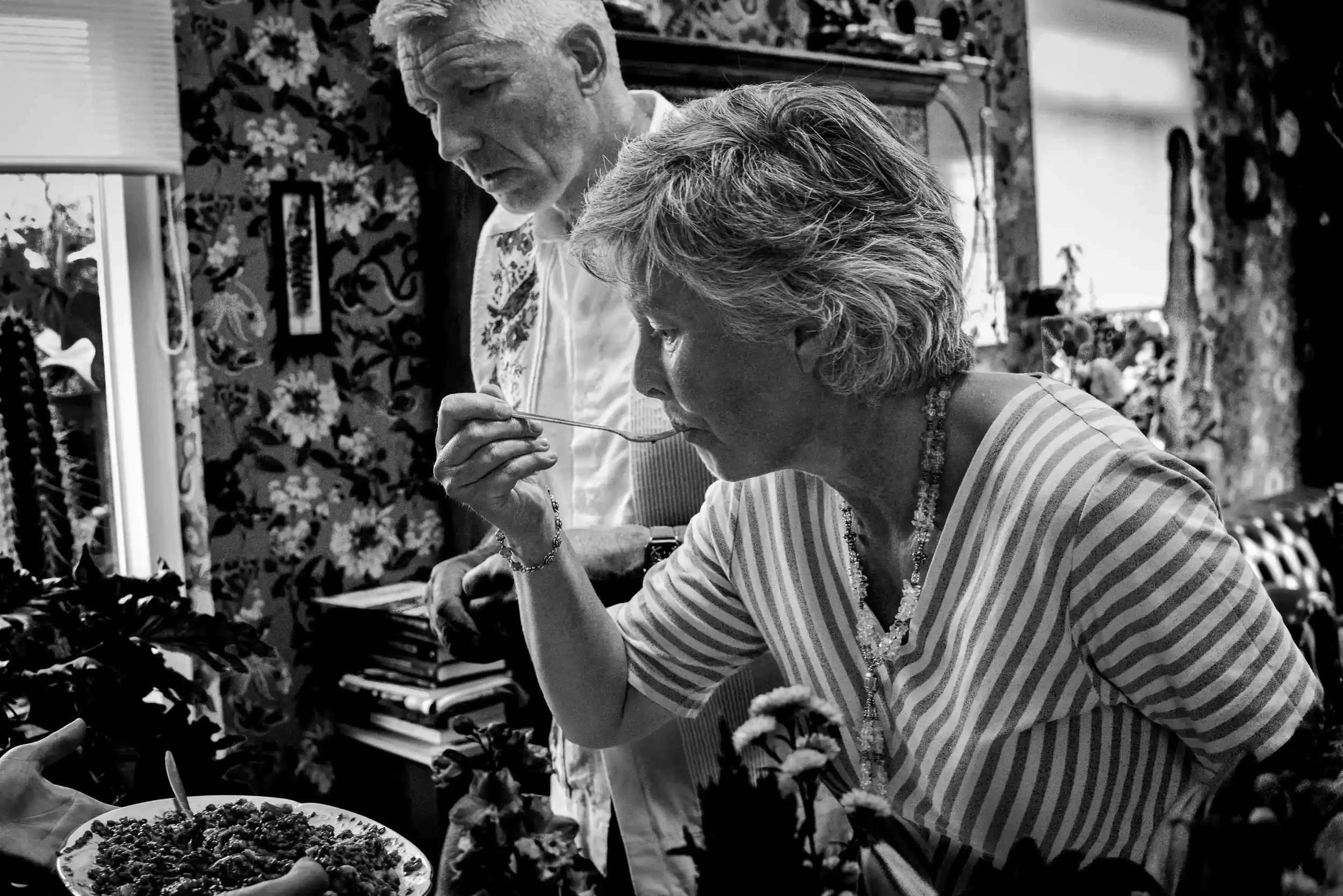 Twee oudere mensen, een man en een vrouw, in een kamer vol bloemen en decoraties, waarbij de vrouw voedzaam voedsel op haar bord eet terwijl de man met een lepel in de hand toekijkt.