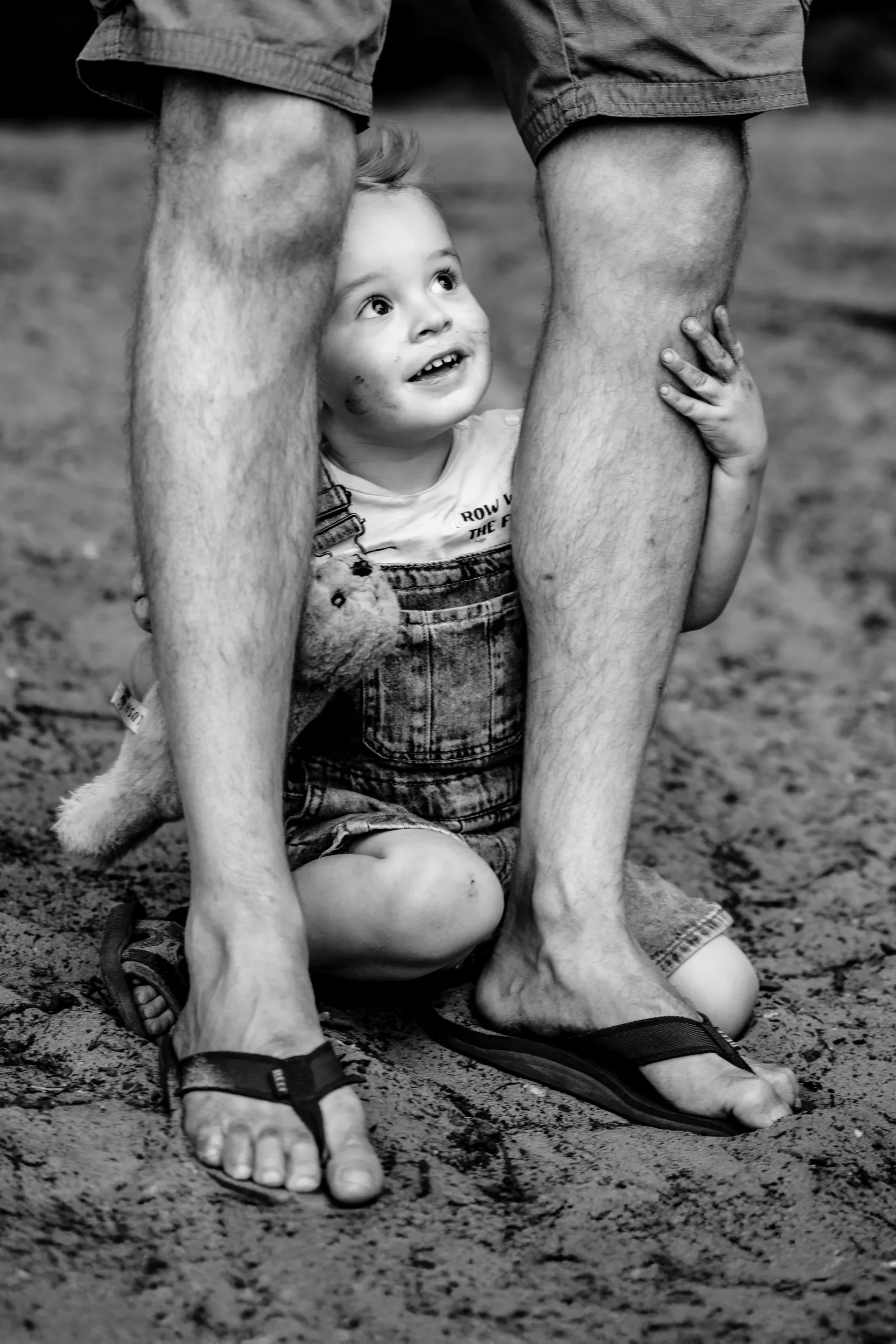 Kleine jongen knielt op zand, omringd door grote volwassenen met benen in korte broek en slippers, kijkt vrolijk omhoog, vasthoudend aan de benen.