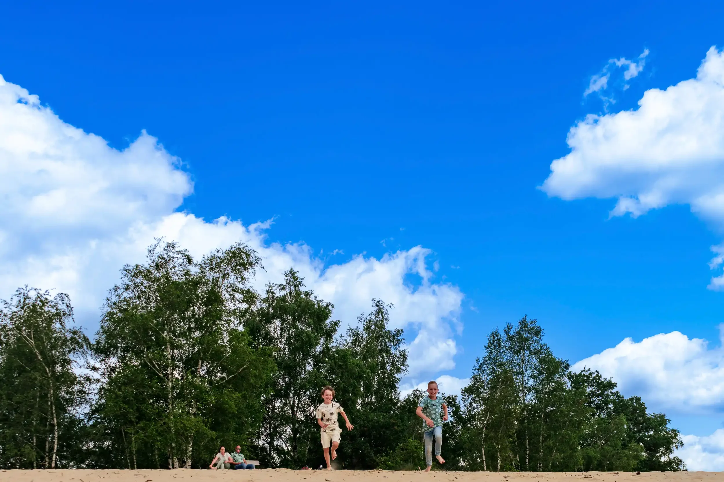 Twee kinderen rennen en lachen op een zandstrand met bomen op de achtergrond onder een blauwe lucht met witte wolken.
