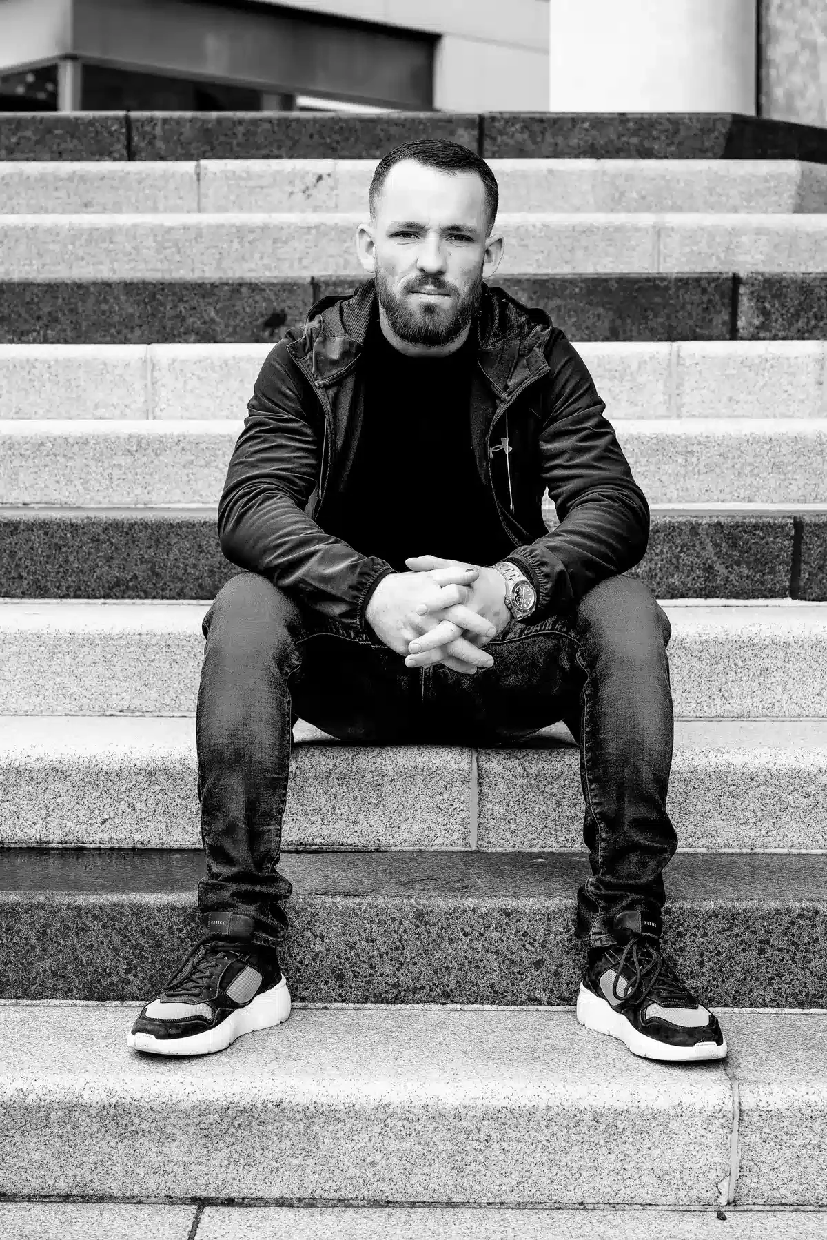 Man zit op trap, gekleed in zwarte jas en broek, draagt sneakers en een horloge, zwart-wit foto.