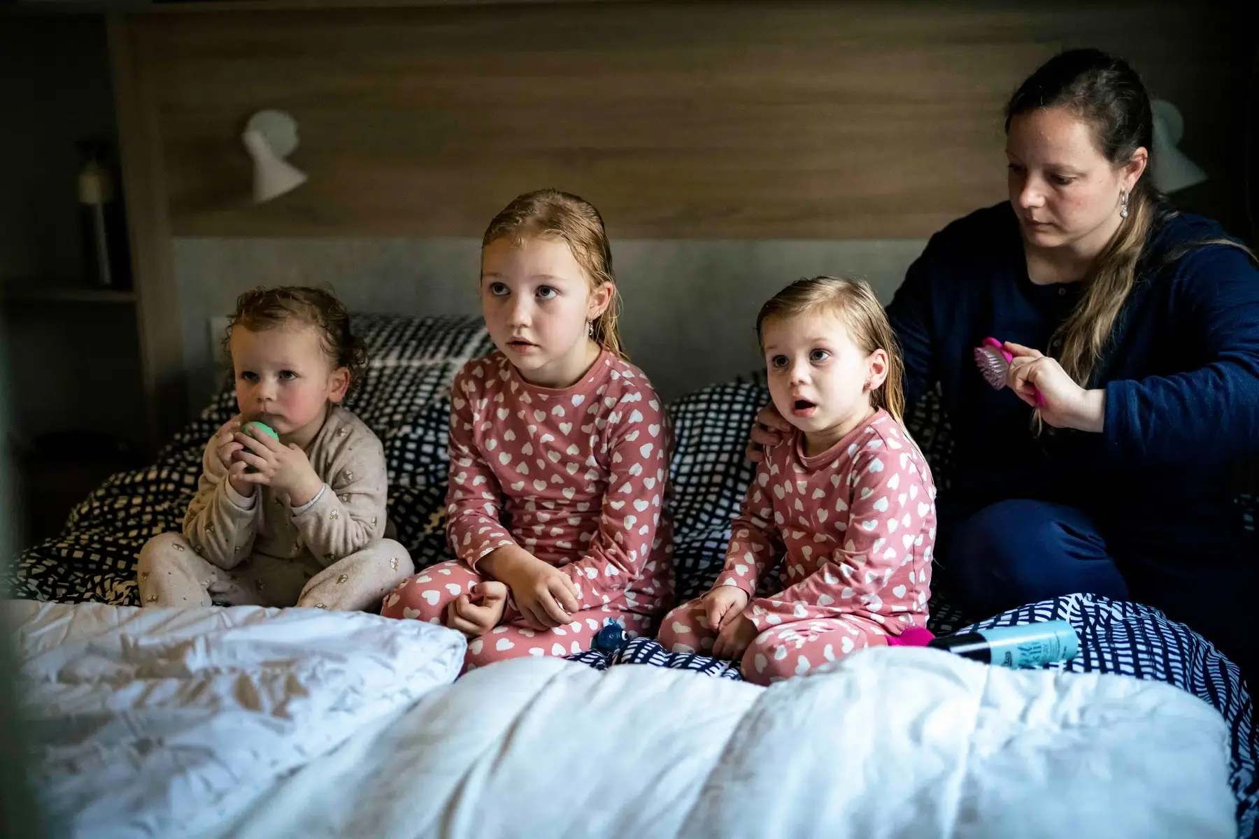 Vier mensen zitten op een bed, drie jonge meisjes in pyjama en een vrouw, terwijl de vrouw haar haar kamt.