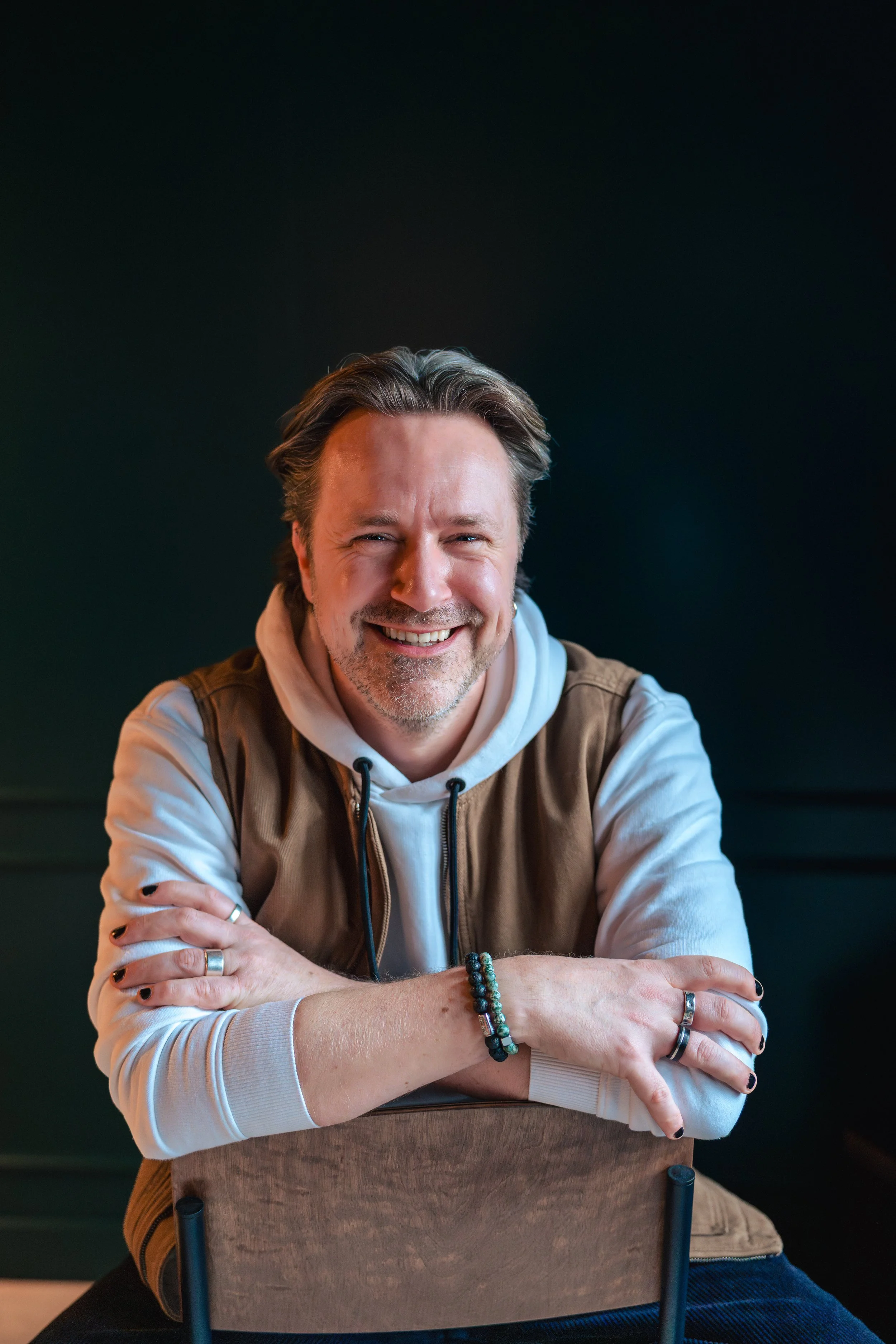 Lachende man zittend achter een houten stoel, met donkere achtergrond.