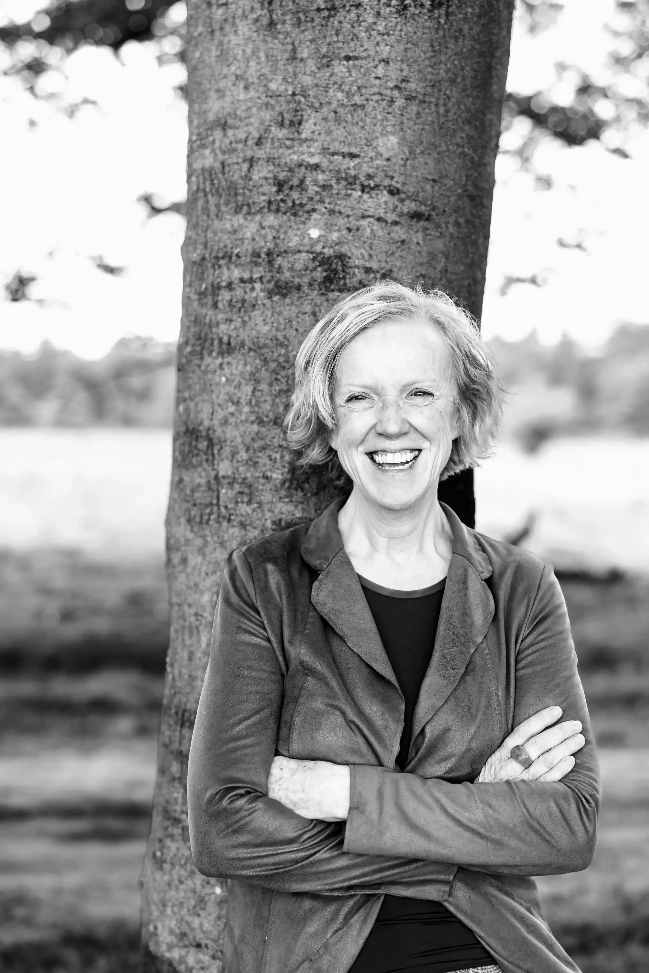 Lachende vrouw in zwart-wit staat voor een boom, armen gekruist, buiten, achtergrond vaag natuur