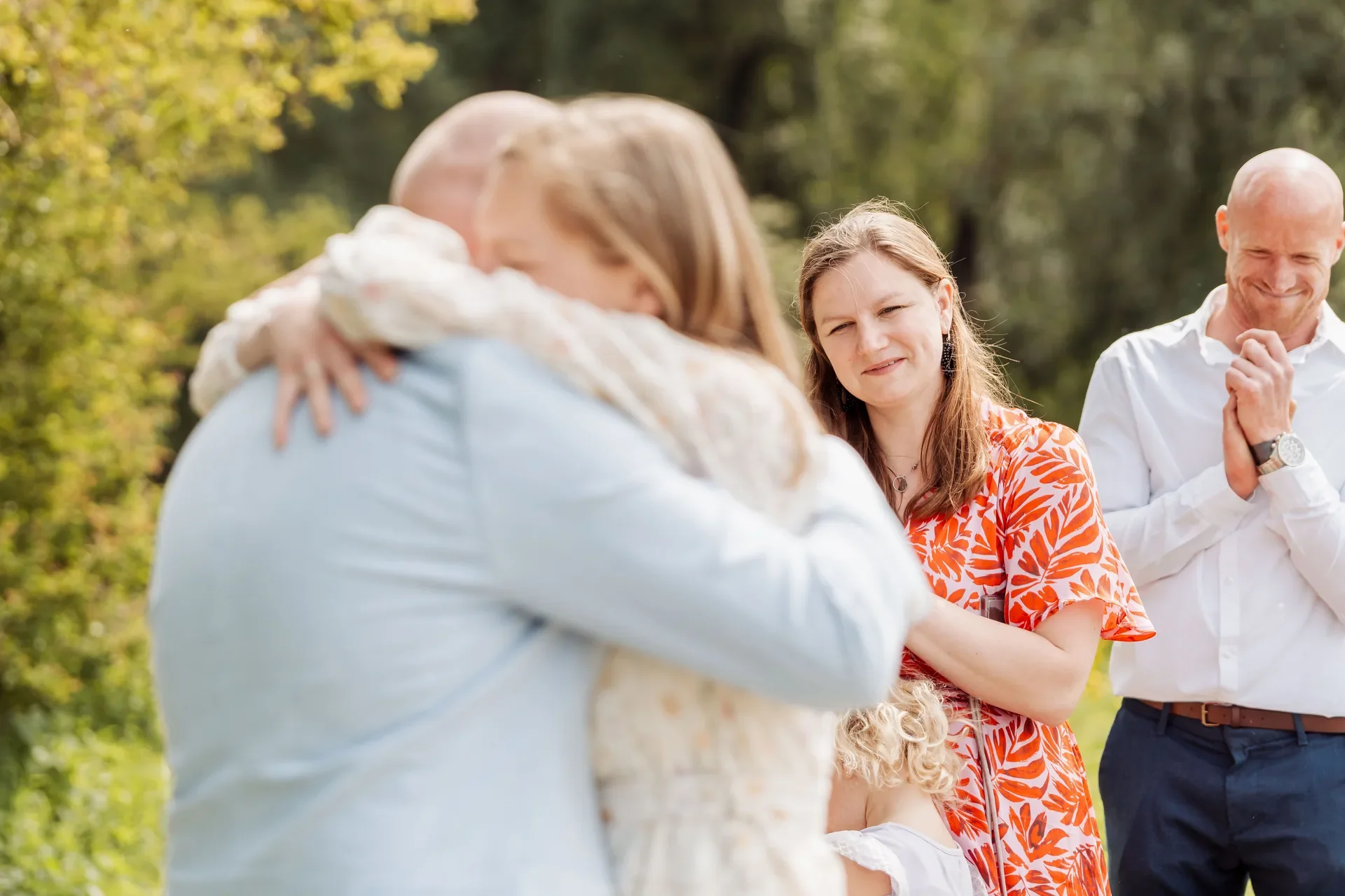 Mensen die elkaar omhelzen tijdens een buitenbijeenkomst in een park, met twee vrouwen en twee mannen die lachen en iemand omhelzen.