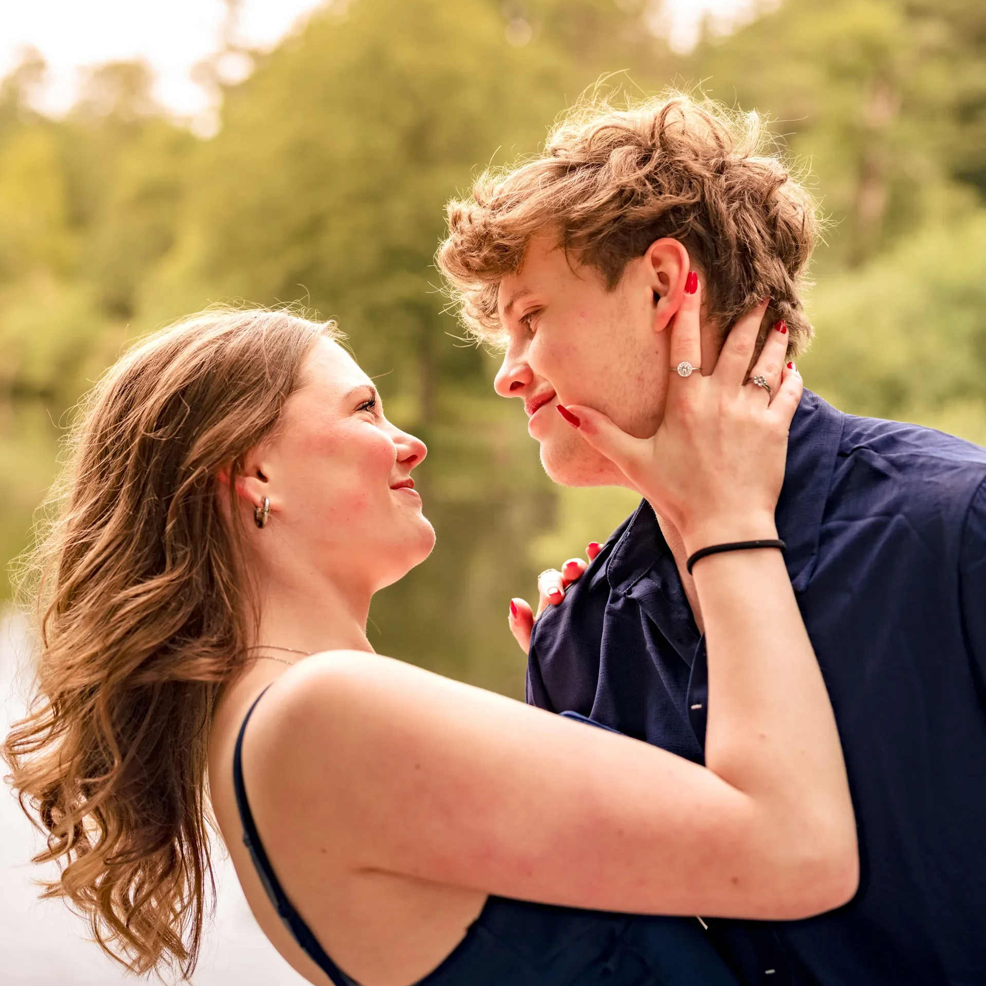 Een vrouw en een man kijken liefdevol naar elkaar, de vrouw houdt de kin van de man vast, ze staan buiten met een boomrijke achtergrond.