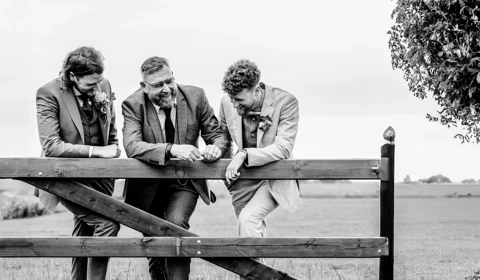 Vier mannen in pakken en blazers lachen en praten samen terwijl ze over een houten hek leunen, buiten op een veld.