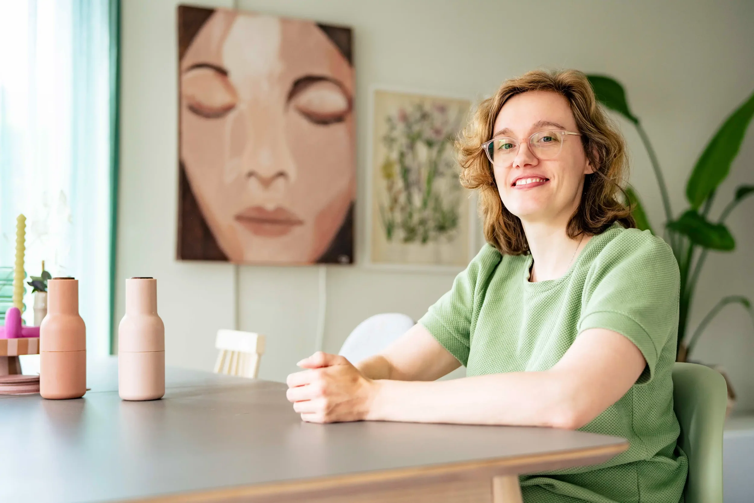 Vrouw met bruin, golvend haar en bril zit aan een tafel in een lichte kamer, met een grote muurdecoratie van een vredige gezichtsuitdrukking en een plant op de achtergrond.