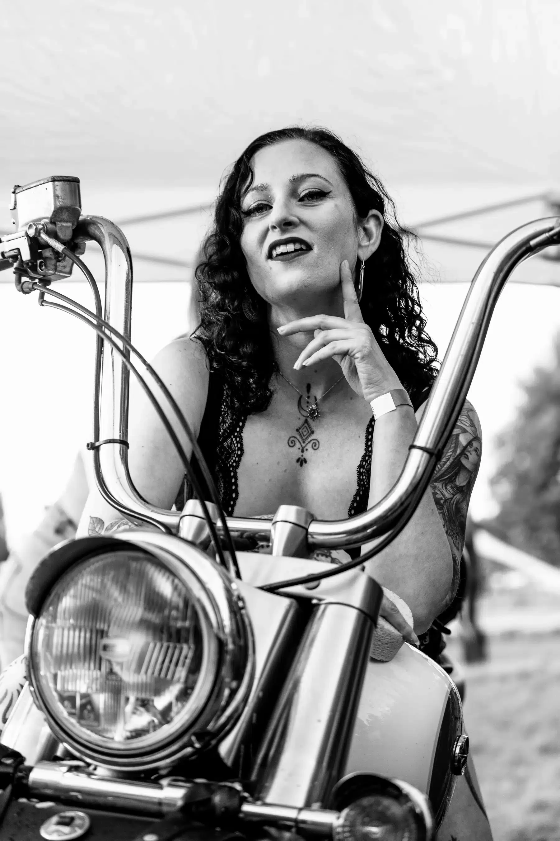 Zwart-wit foto van een vrouw met donkere krullen op een motorfiets, met een sierlijke tattoo op haar linkerarm, wijzend naar haar gezicht met een glimlach en een vinger op haar wang.