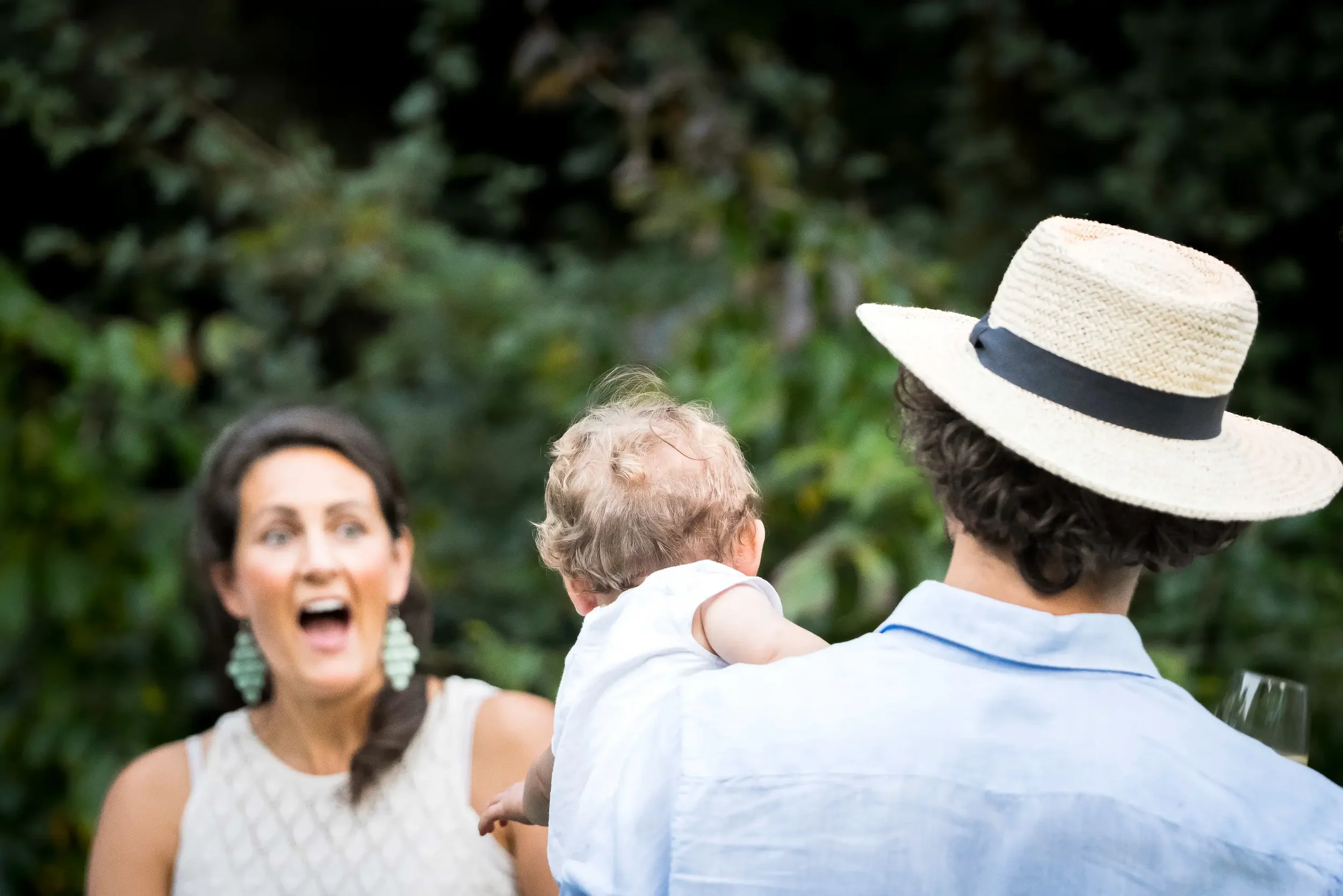 Een vrouw reageert verbaasd of geschrokken terwijl ze een man en kind ziet, die van achteren worden gefotografeerd buiten. De man draagt een strooien hoed en een lichtblauw overhemd, en houdt een glas wijn vast. Het kind zit op zijn schouder.