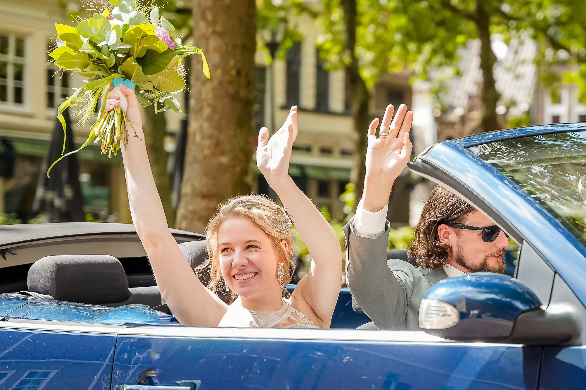Twee mensen rijden in een blauwe cabrioletauto. De vrouw zwaait met een bloemenbundel en lacht. De man zit naast haar en kijkt naar voren. Ze zijn buiten op een straat met bomen en gebouwen.