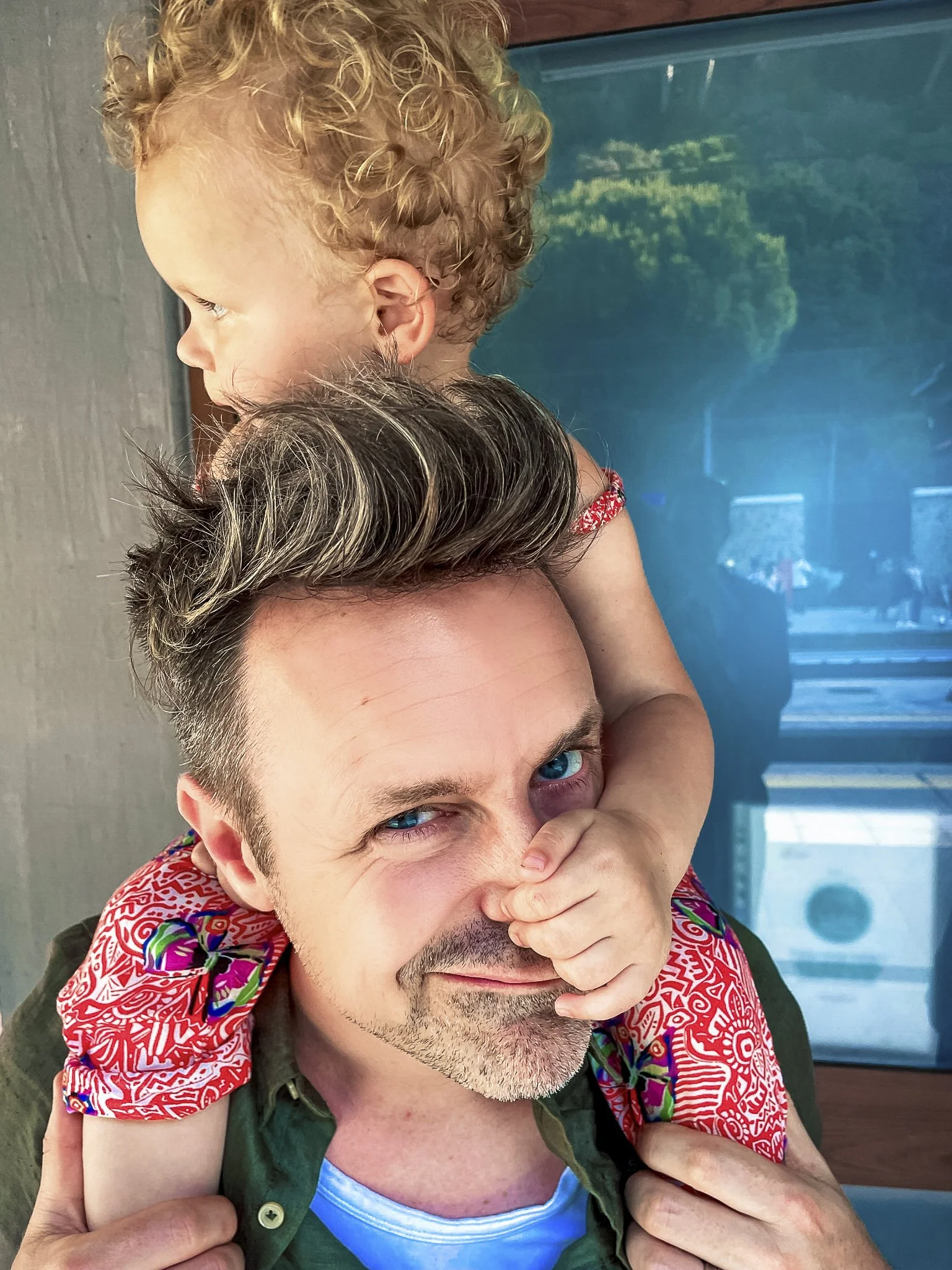 Man met kort, grijzend haar en blauwe ogen met een kind op zijn schouders, dat een rode bandana en een kleurrijke blouse draagt, beide lachen, met een blauwkleurige achtergrond.