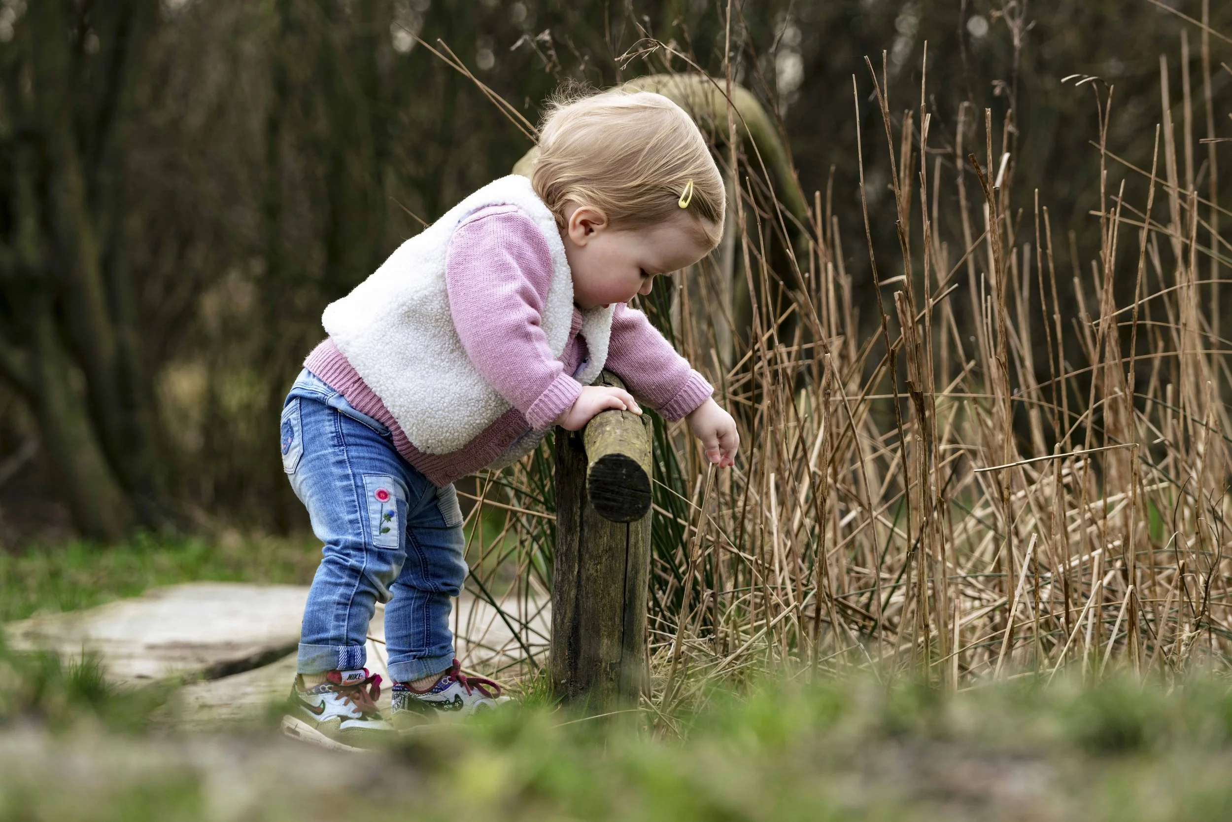 Een jonge jongen die op een houten pad in een natuurlijke omgeving staat, terwijl hij zich naar een ander stuk natuur buigt en naar iets op de grond kijkt.