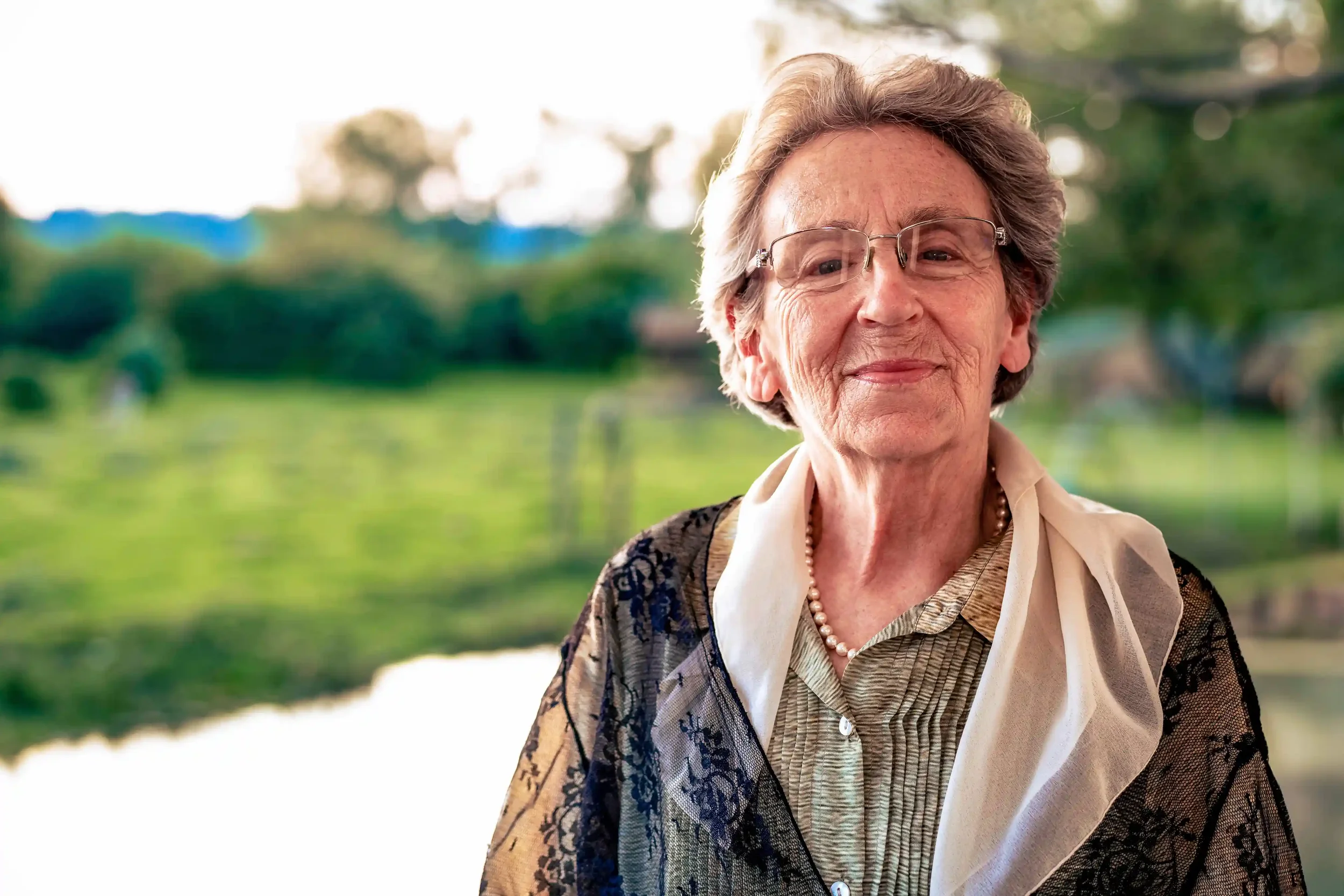Een oudere vrouw met grijs haar en brillen, glimlachend, gekleed in een blouse, een parelketting en een zwarte kanten jas, staat buiten met een groene landschap op de achtergrond.