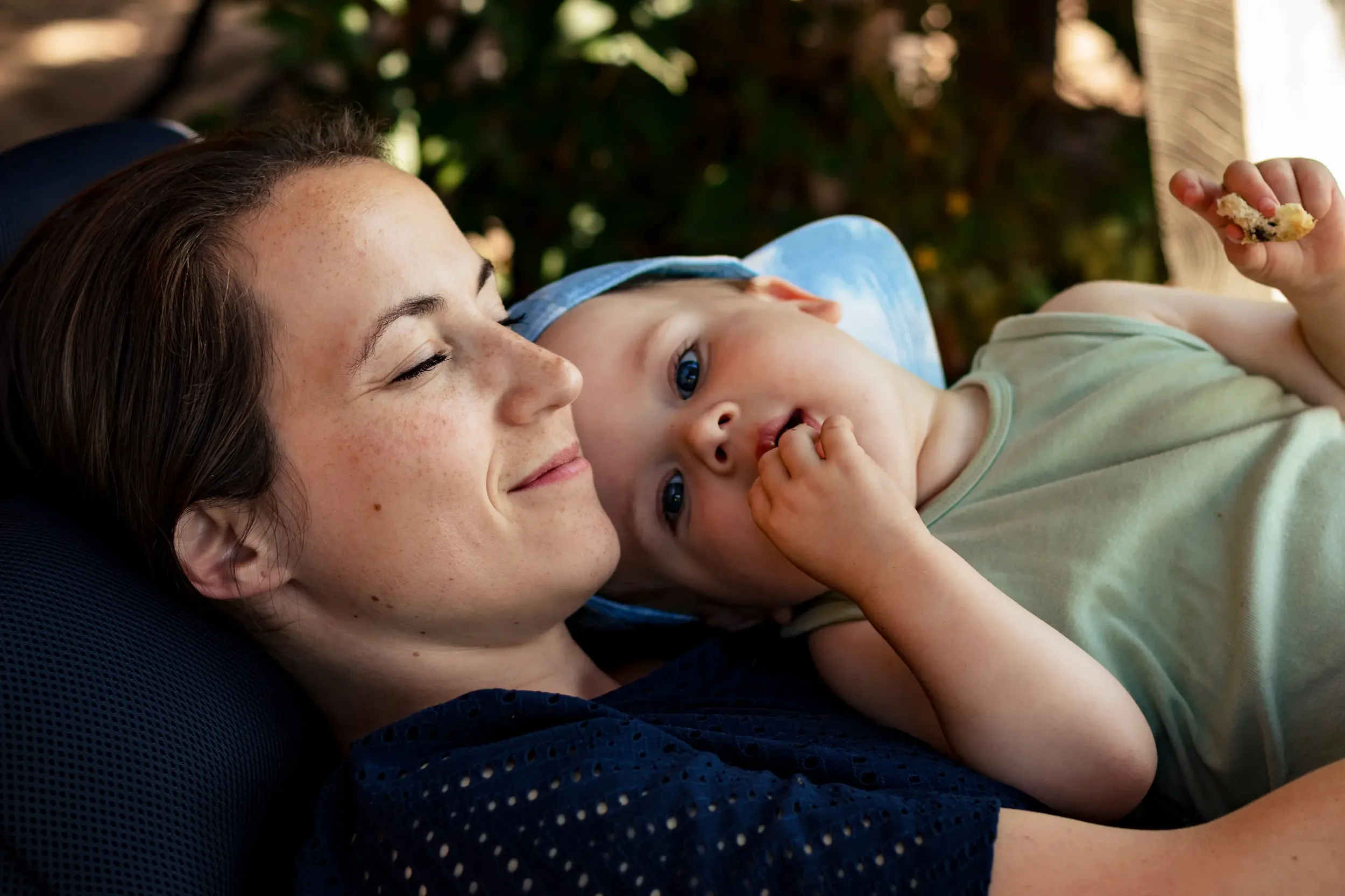 Een jonge vrouw ligt buiten op een bankje, knuffelt een slapende peuter met een blauwe pet. De peuter eet een stukje koekje en kijkt naar de camera, terwijl de vrouw glimlacht en haar ogen gesloten heeft.
