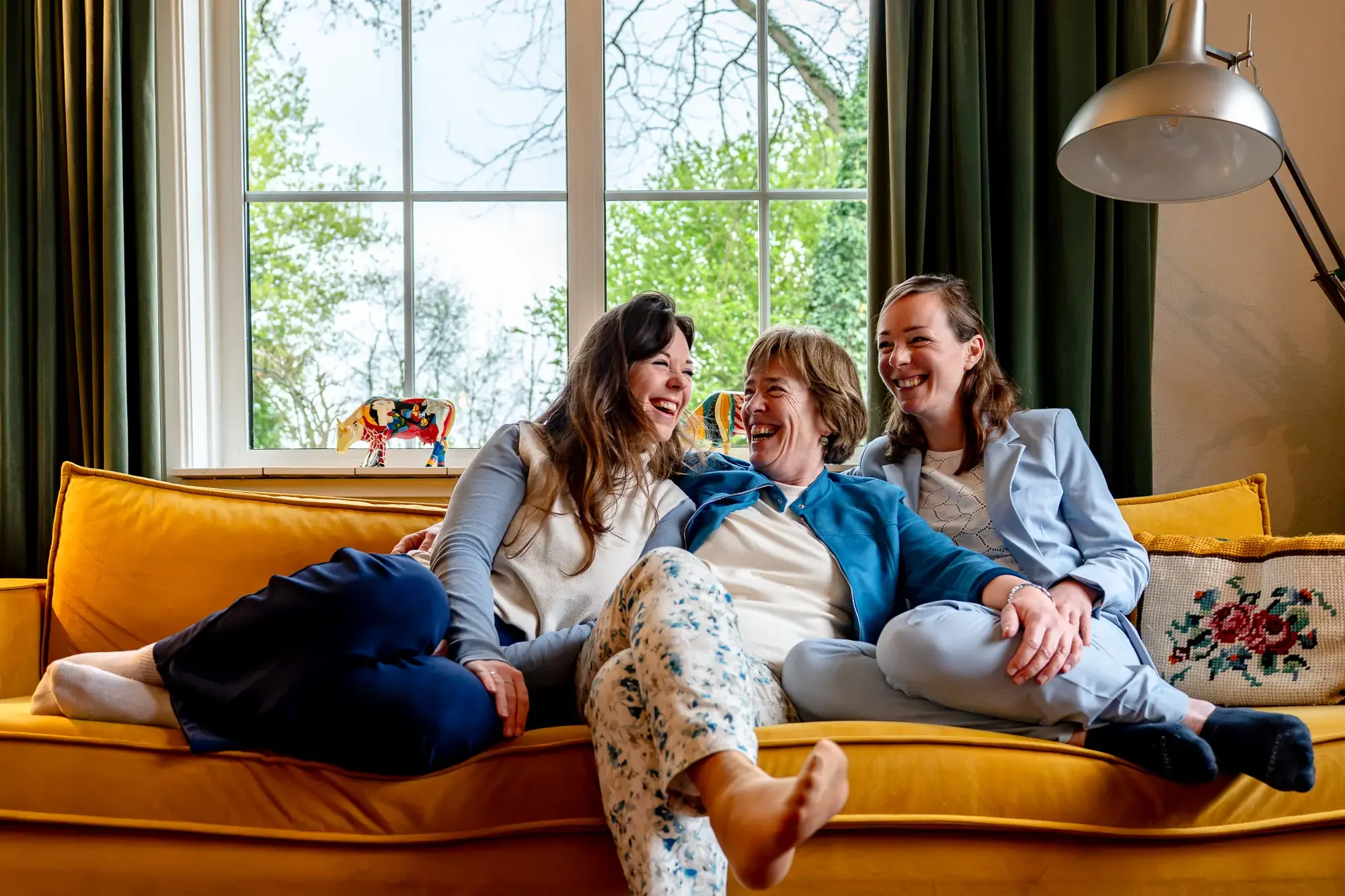 Vijf mensen zitten op een gele bank in een woonkamer, lachen en hebben plezier. Achter hen grote ramen met groen uitzicht.