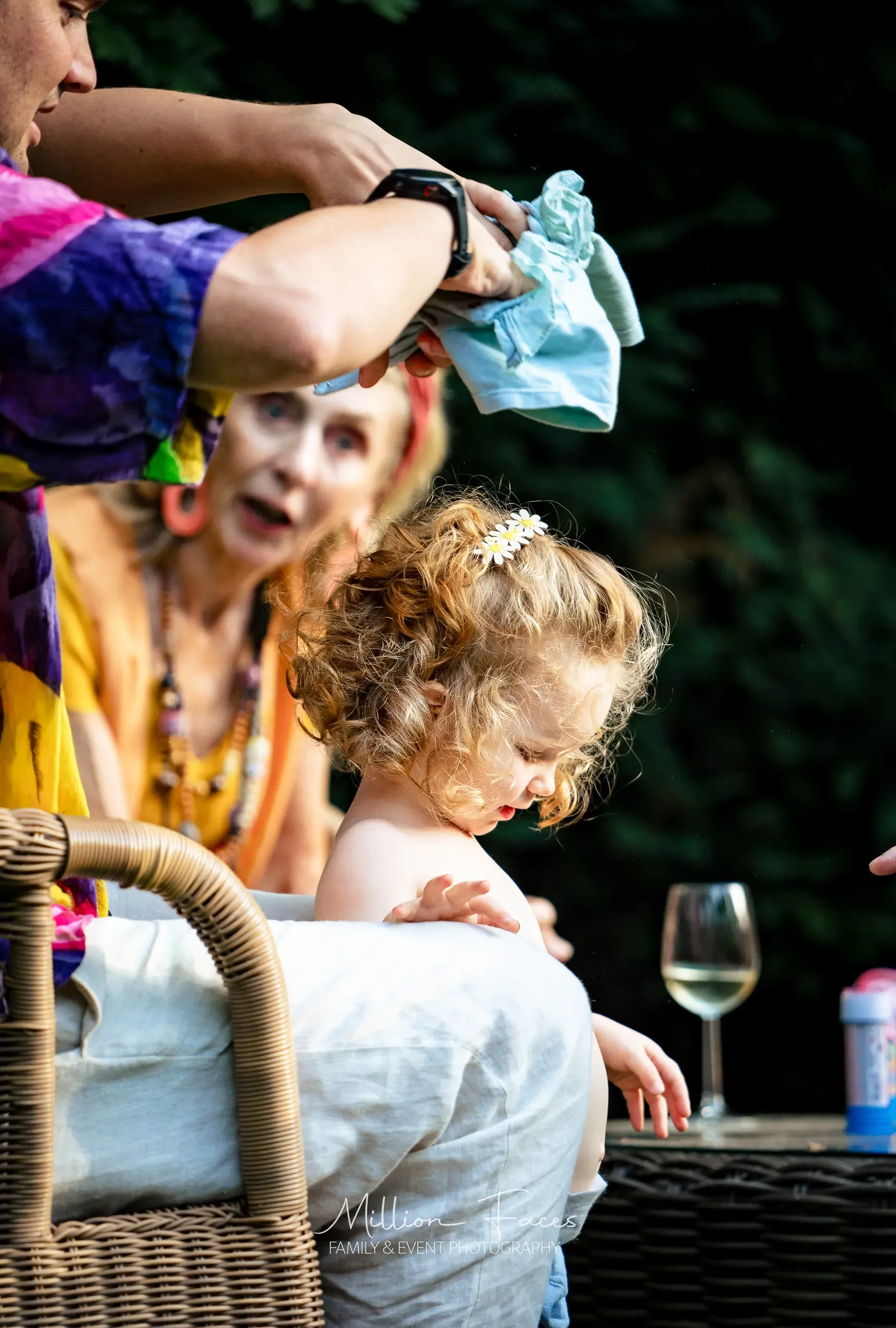 Een jonge meisje met krullend lichtbruin haar zit op een bank, terwijl een vrouw haar een kledingstuk geeft. Een andere vrouw in de achtergrond kijkt verbaasd. Er staat een glas witte wijn op een tafel naast de bank.