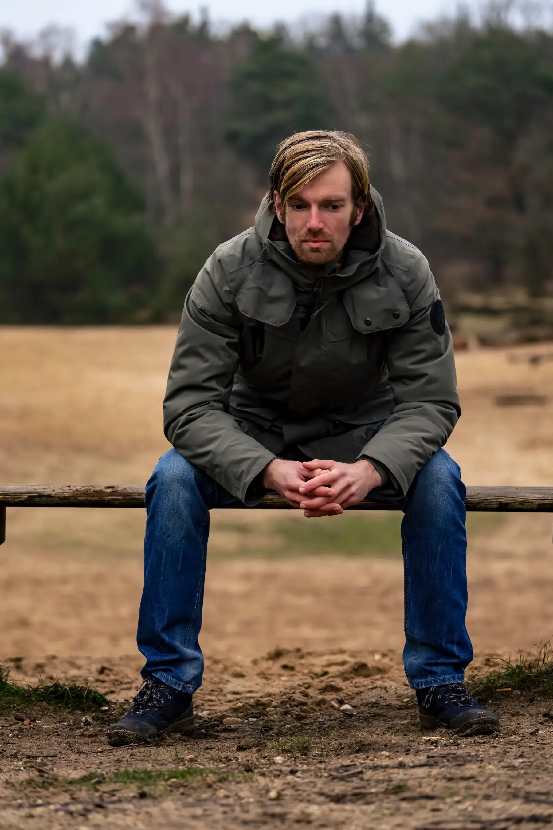 Man zit op een houten bankje in een open veld, omgeven door bomen, met een rustige uitdrukking.