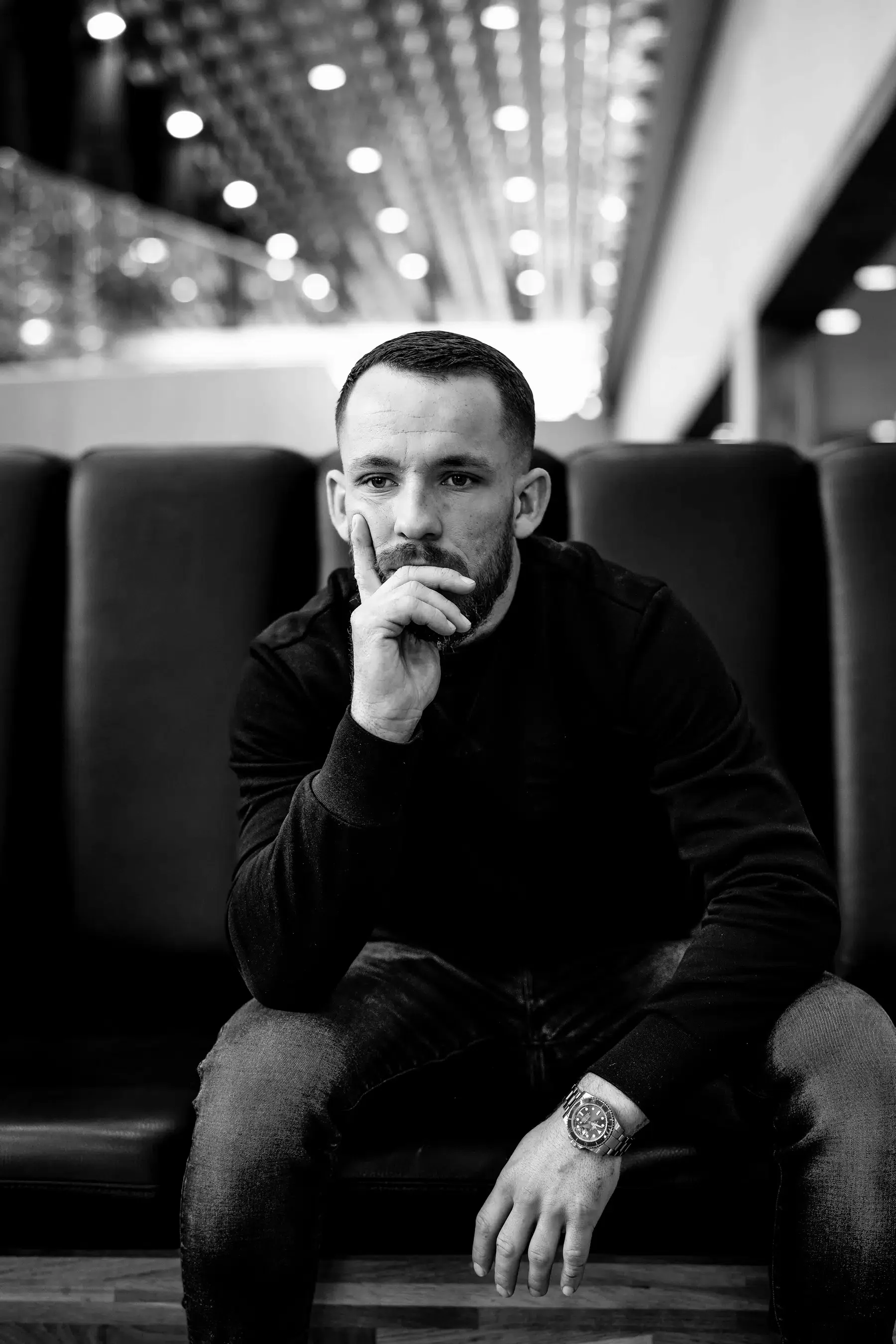 Zwart-wit foto van een man die op een bank zit, met zijn hand op zijn kaak, in een interieur met een plafond vol lampen.