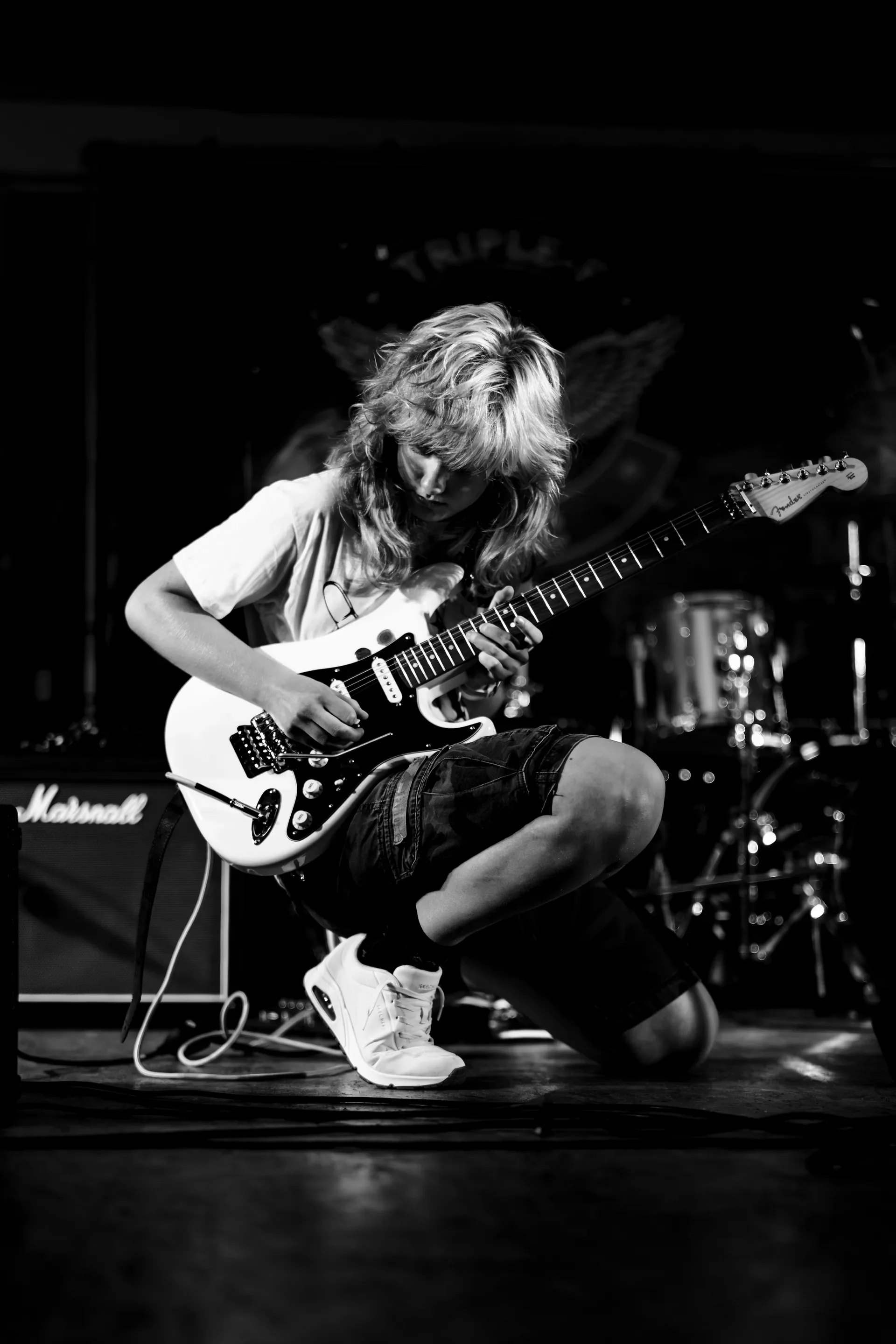 Een jonge persoon met lang haar speelt gitaar op een podium, omgeven door muziekapparatuur, in zwart-wit.