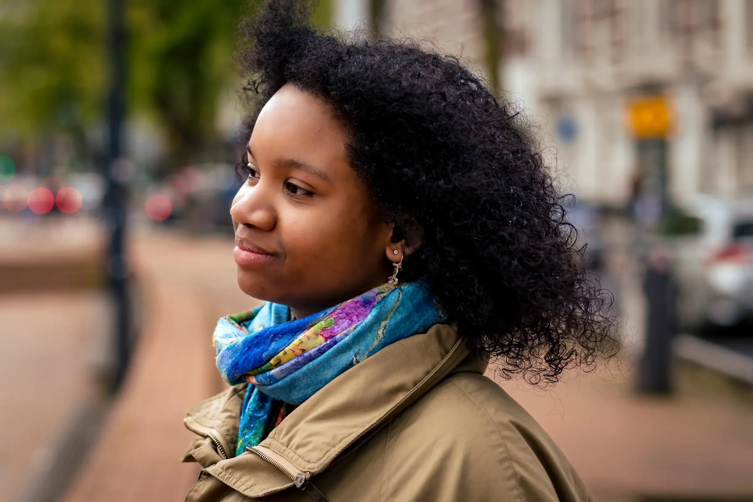 Portret van een jonge vrouw met dik, curly zwart haar, een kleurrijke sjaal en een beige jas, staand op een straat met bomen en geparkeerde auto's op de achtergrond.