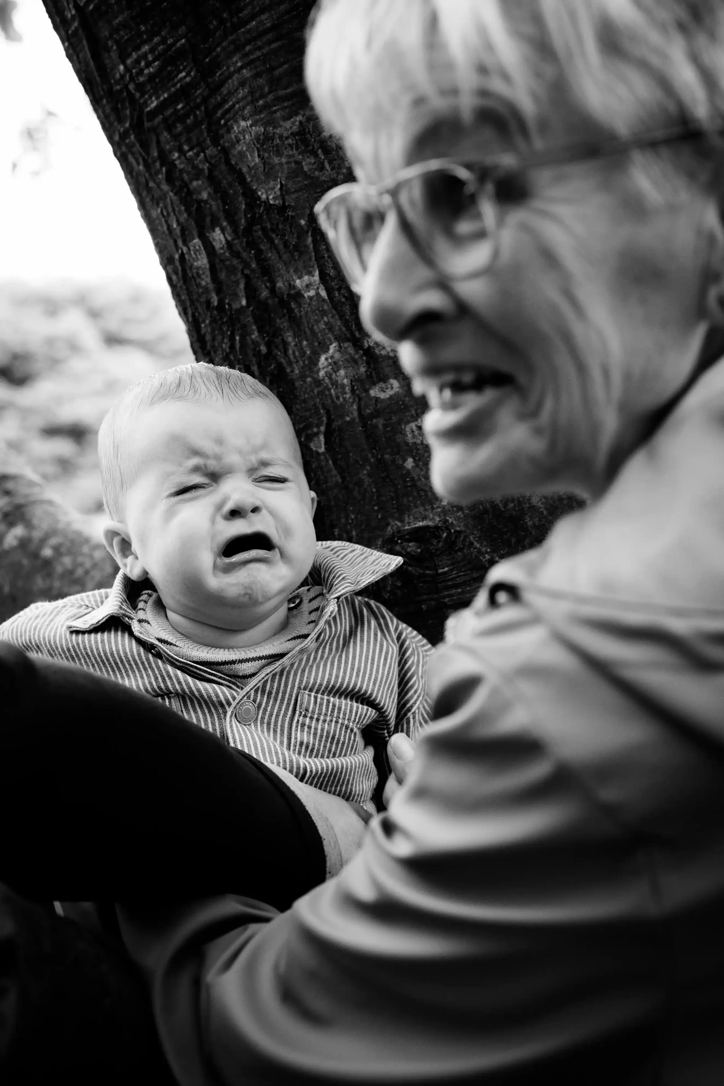 Een close-up van een huilende baby met een oudere vrouw die met haar spreekt, buiten bij een boom.