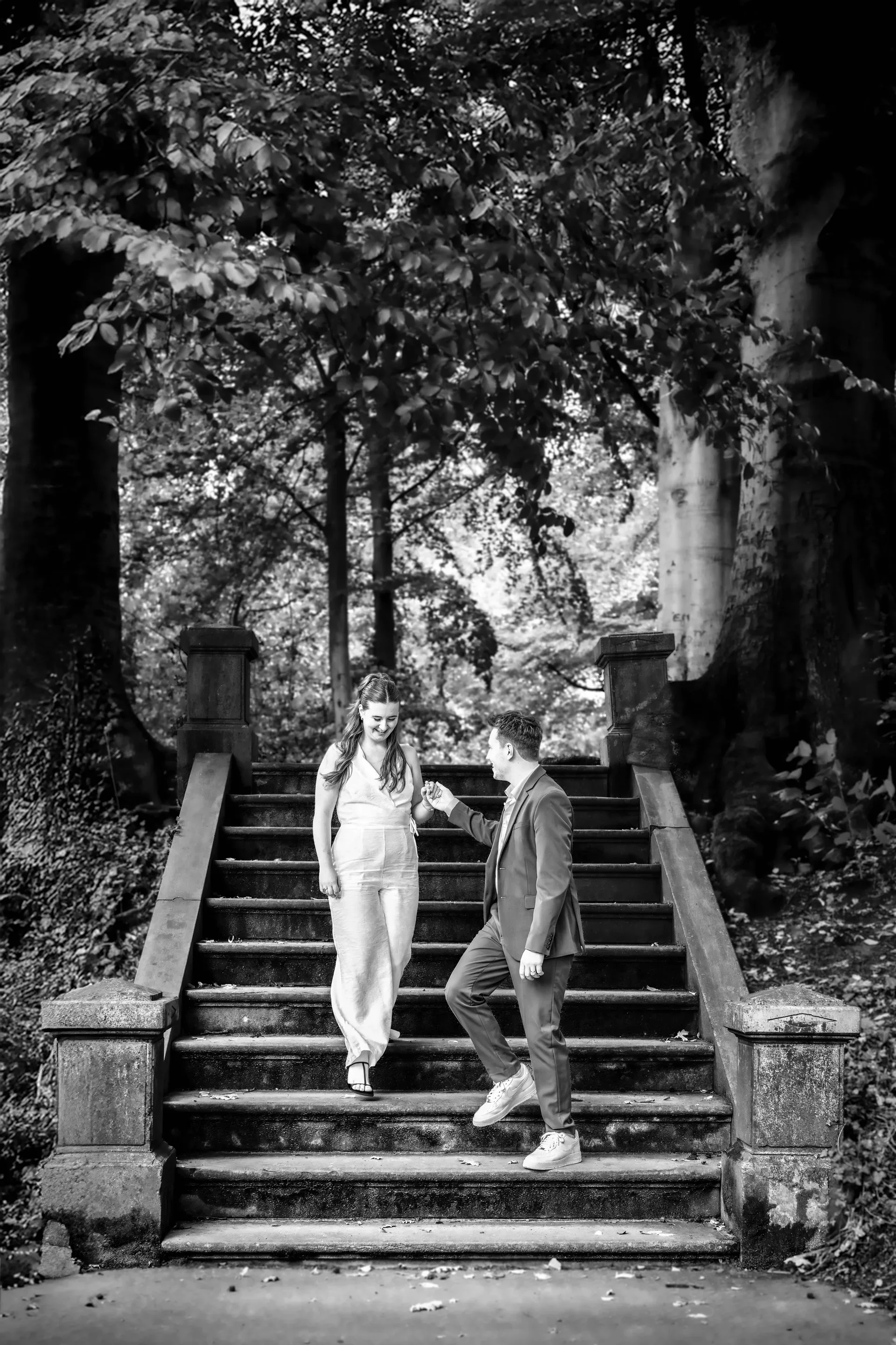 Een man en een vrouw staan op een stenen trap in een bos, waarbij de man de vrouw iets geeft of overhandigt. De afbeelding is in zwart-wit.