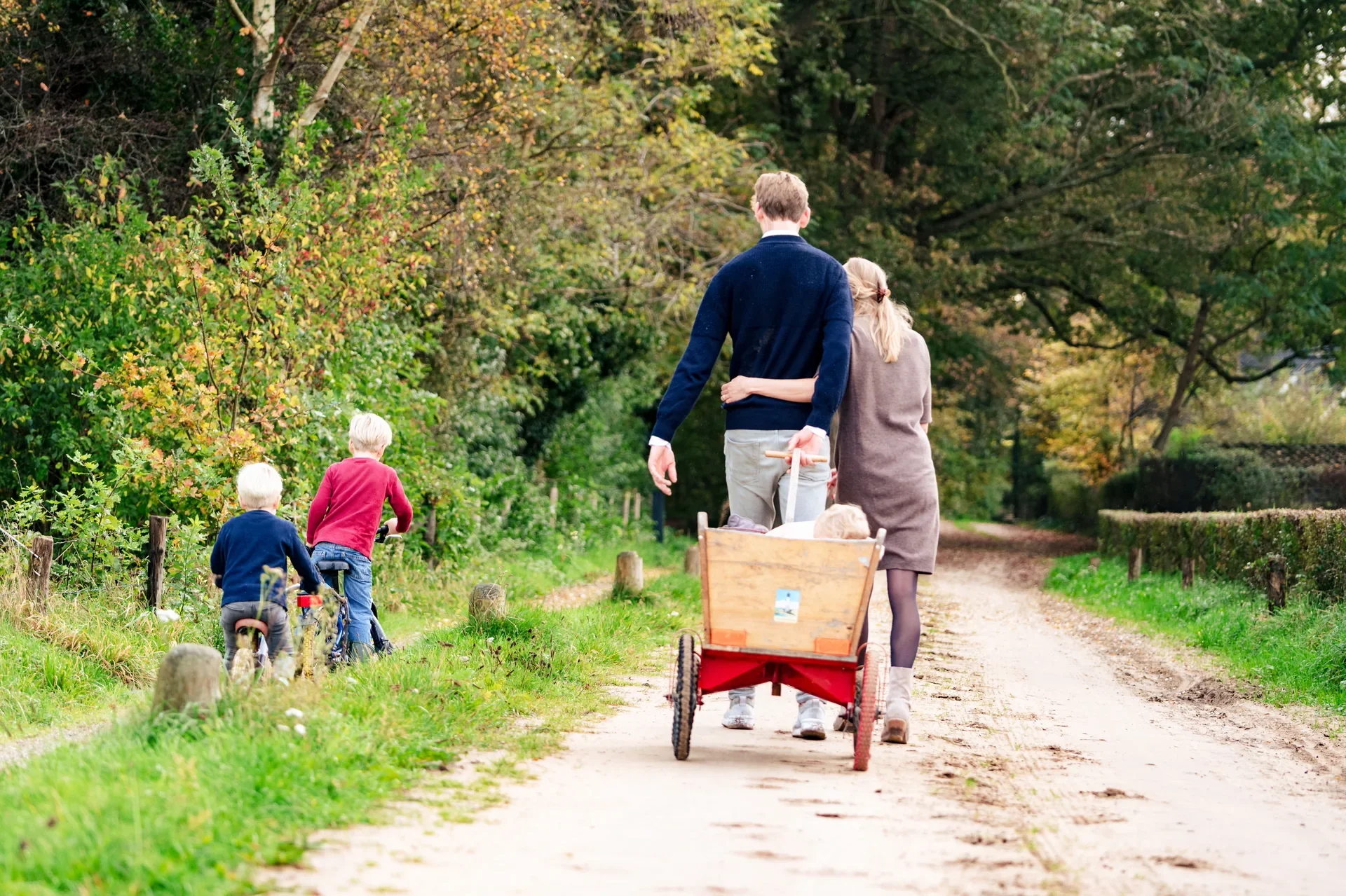 Een gezin wandelt op een bospad in de herfst, met twee kinderen op fietsen en volwassen met een kinderwagen vol spullen.