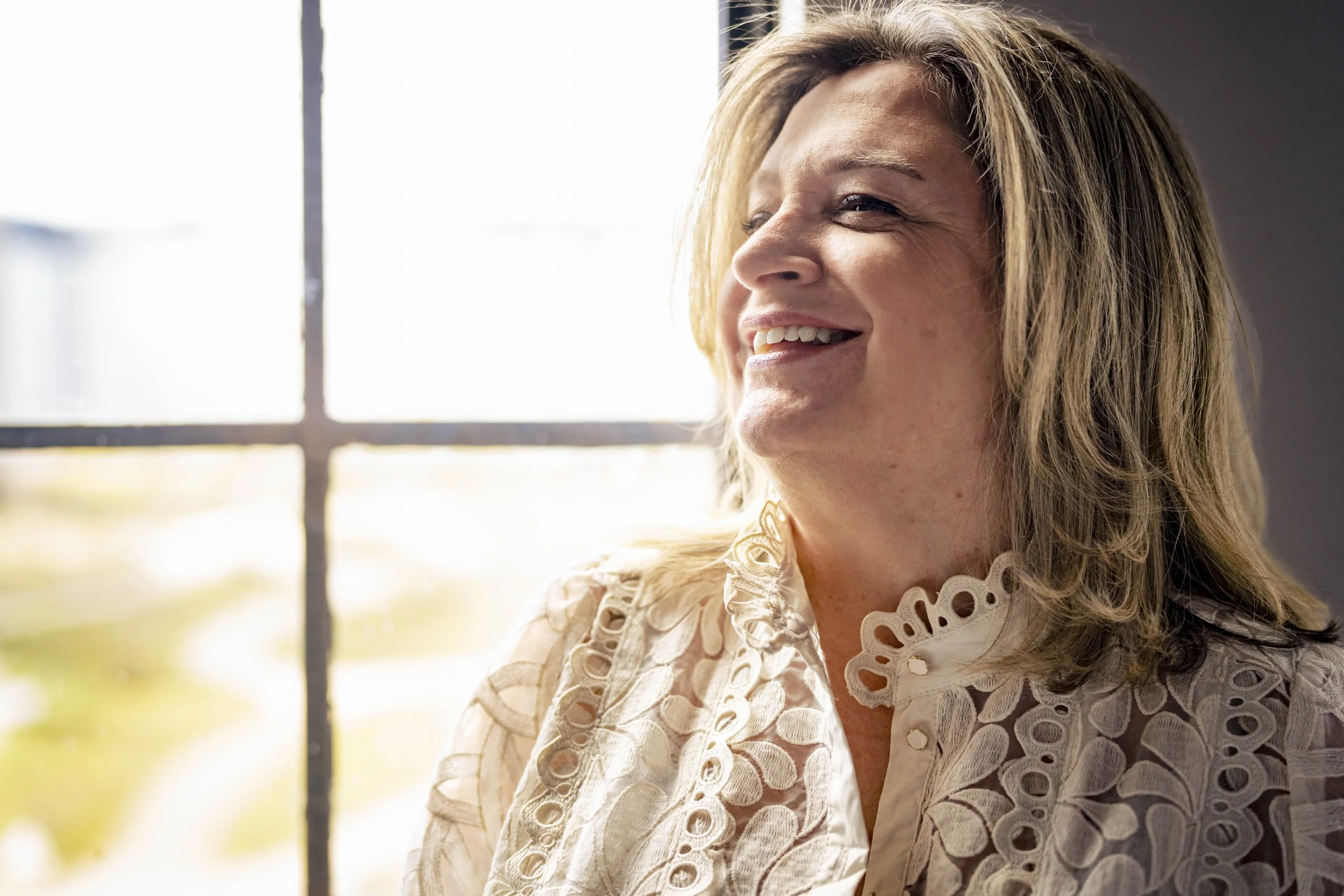 Close-up van een lachende vrouw met blond haar die naar buiten kijkt naast een raam met een kruisvormige raamstijl, door het raam schijnt fel zonlicht.