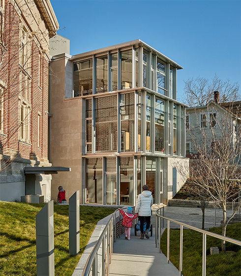 People walking on a pathway in front of a modern glass building with multiple floors, surrounded by grass and trees, under a clear blue sky.