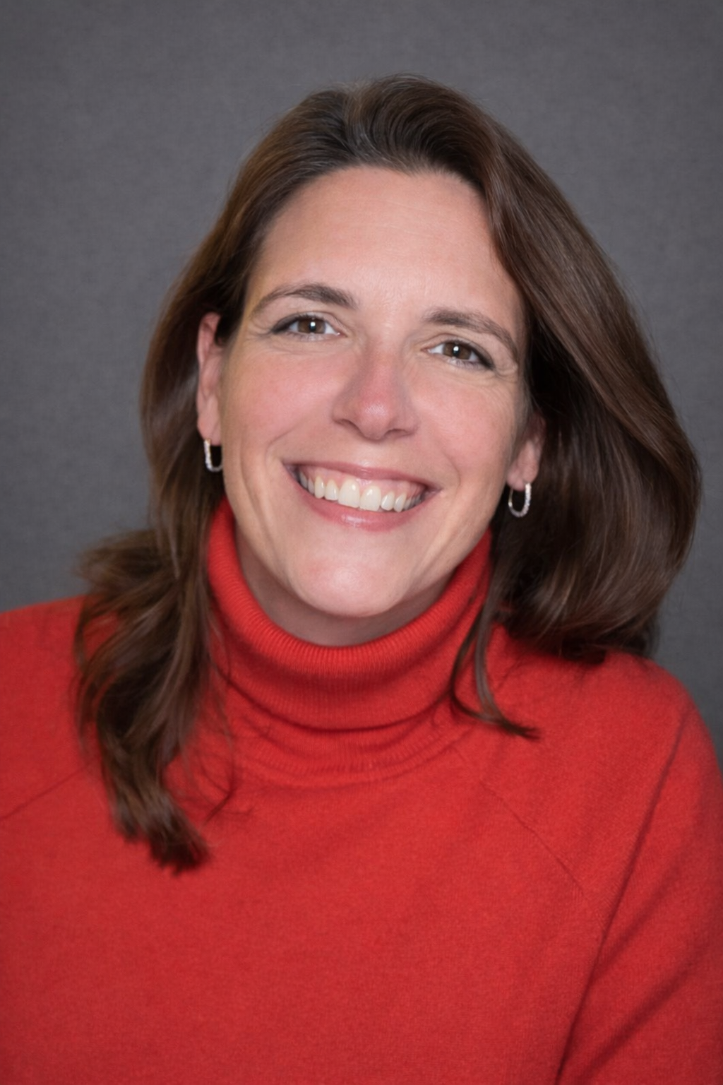 A woman with shoulder-length brown hair, wearing a red turtleneck sweater, smiling at the camera against a gray background.