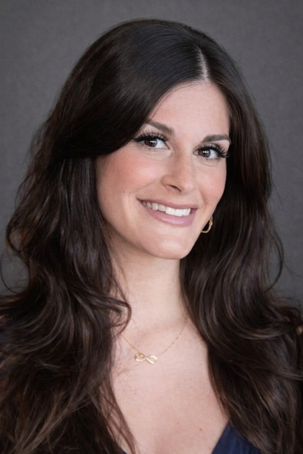 A woman with long, wavy dark brown hair, smiling with white teeth, wearing a black top, gold infinity necklace, and small gold hoop earrings, against a plain gray background.
