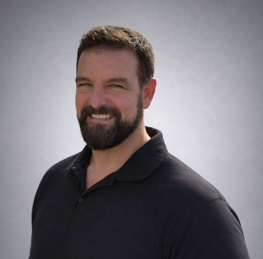 A man with short brown hair, a full beard, and a mustache, smiling and wearing a black polo shirt, standing against a light gray background.