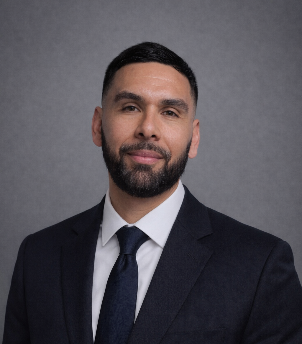 Professional headshot of a man with dark hair and a beard, wearing a black suit, white shirt, and black tie, against a gray background.