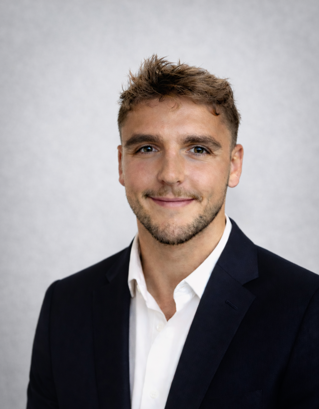 A young man with short, tousled light brown hair, blue eyes, and a light beard, dressed in a dark blazer and white shirt, smiling against a plain light gray background.
