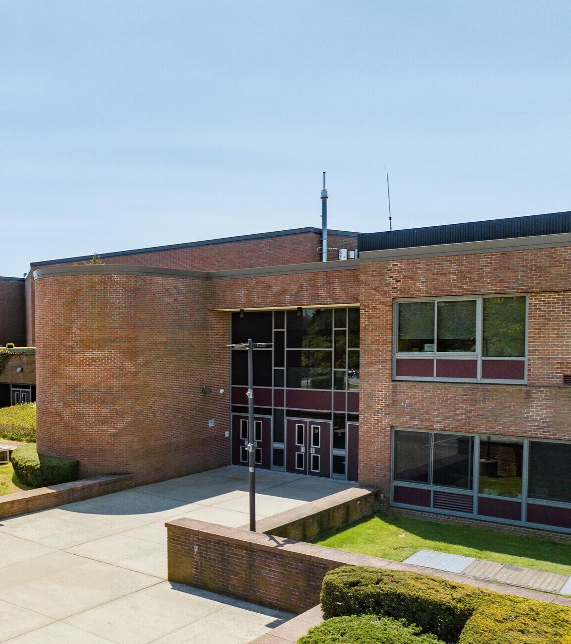 A brick school building with large windows and a main entrance, surrounded by a paved courtyard, bushes, and grassy areas, under a clear blue sky.