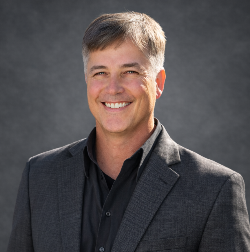 A smiling man with short gray hair wearing a dark suit and black shirt, standing against a gray background.