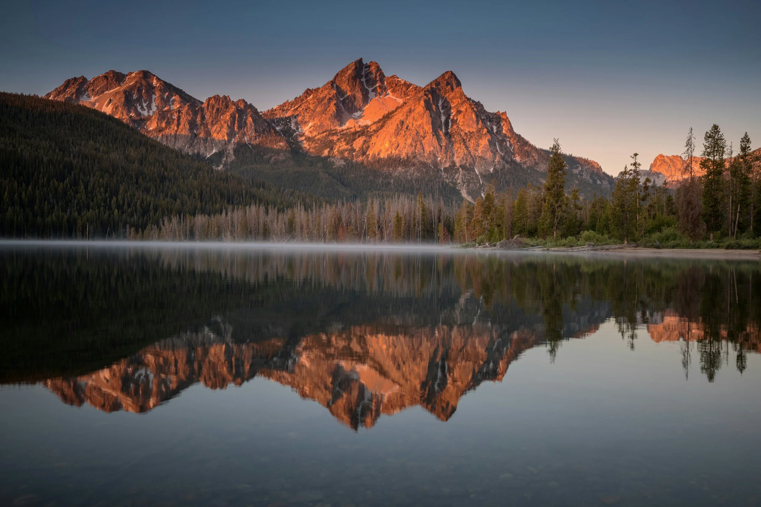 Sunset illuminating rugged mountain peaks over a calm lake, with trees along the shoreline and their reflection in the water.