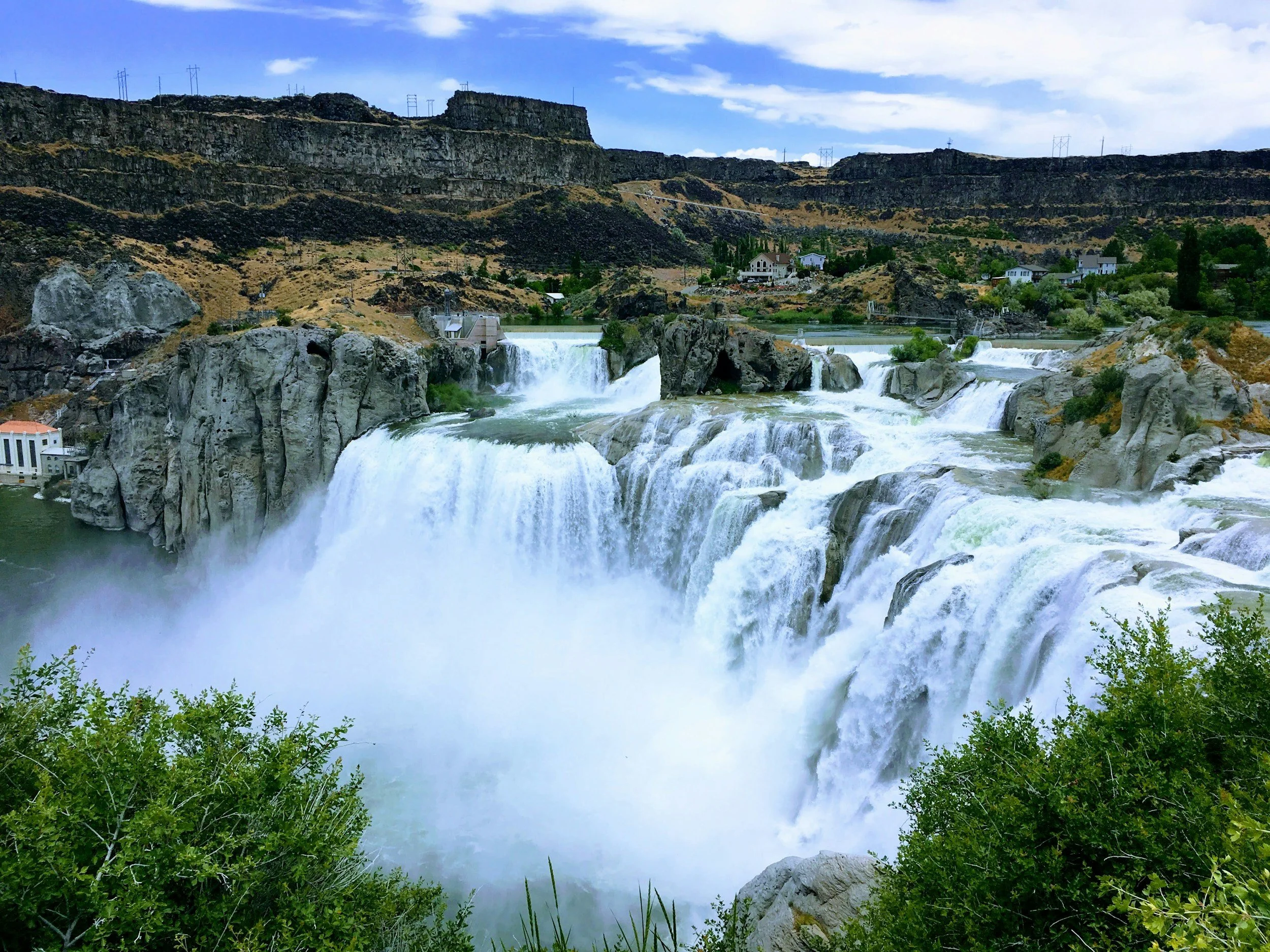 Waterfall cascading over rocks with small houses and green trees on the hillside in the background under a partly cloudy sky.