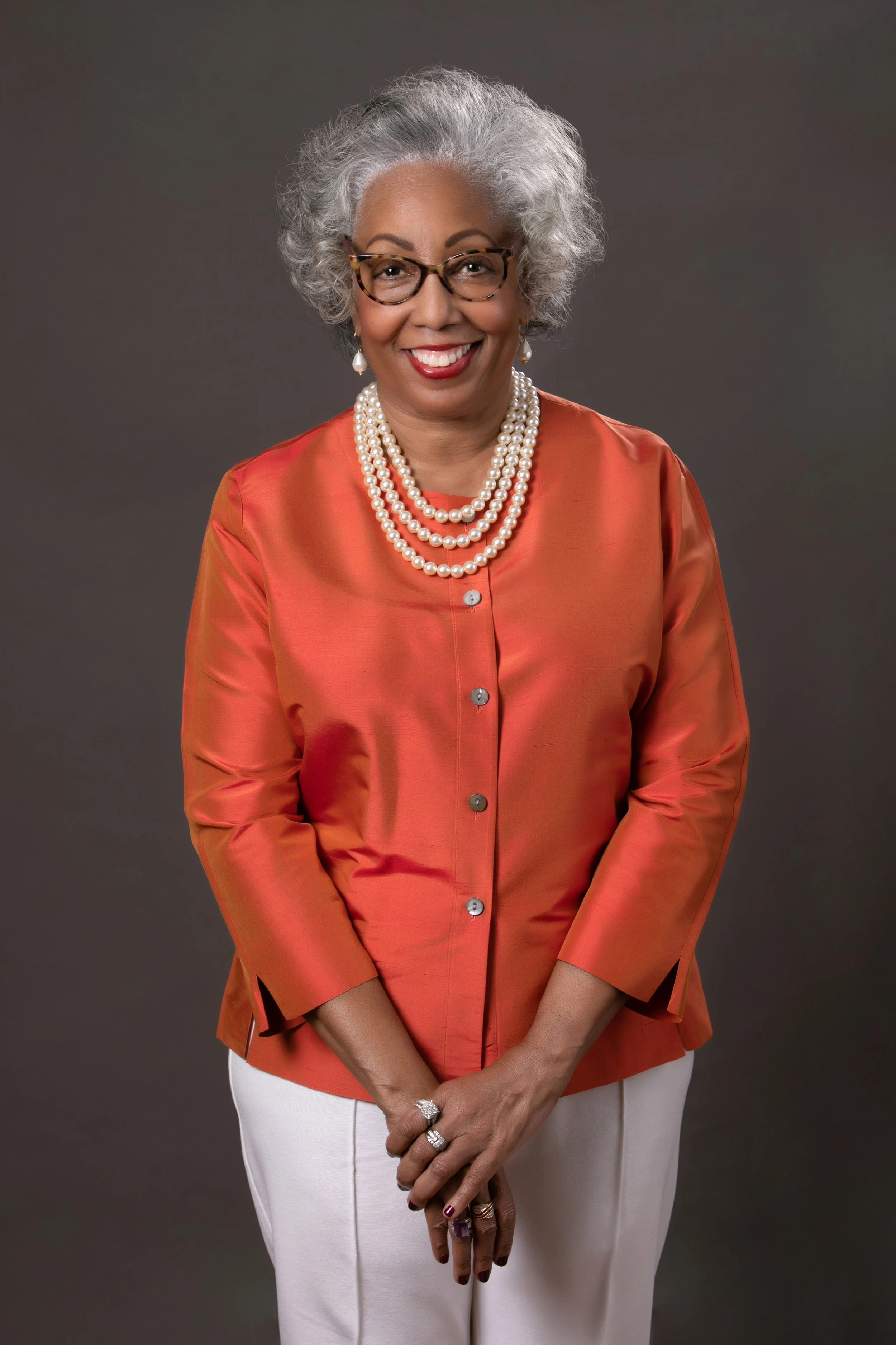 Photo of Dr. Sandra Tanner, author. A beautiful black woman with salt and pepper hair. She wears a bright orange top and white pants with a three strand pearl necklace. She has a huge smile.