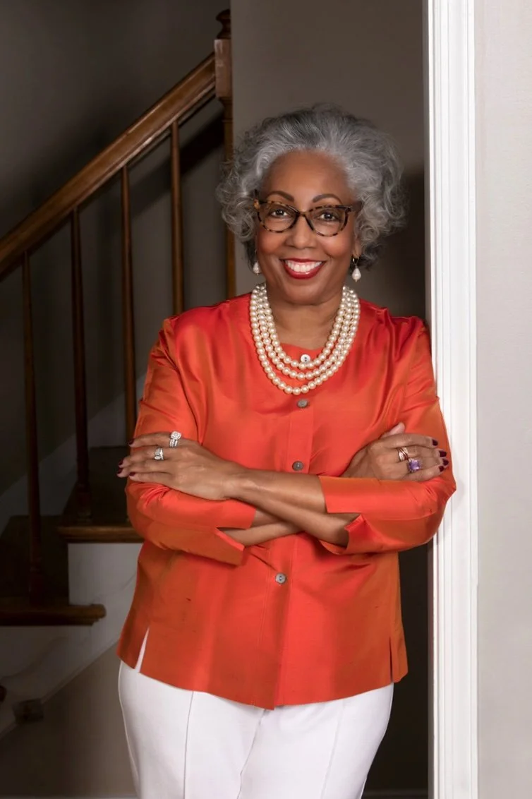 Photo of Dr. Sandra Tanner, author. A beautiful black woman with salt and pepper hair leaning against a doorway. Her arms are casually folded.. She wears a bright orange top and white pants with a three strand pearl necklace. She has a huge smile.