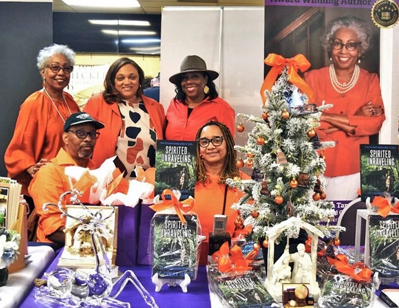 A group of people stand behind a display table at a book fair. A Christmas tree is on thetable along with several copies of the book Spirited Unraveling.