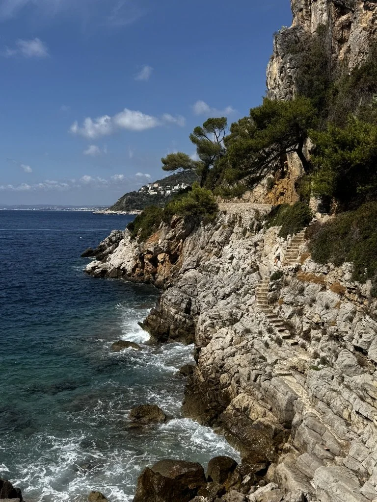 A rocky coastline with stairs built into the rock cliffside. Green trees grow on the cliff, overlooking the blue ocean under a partly cloudy sky.