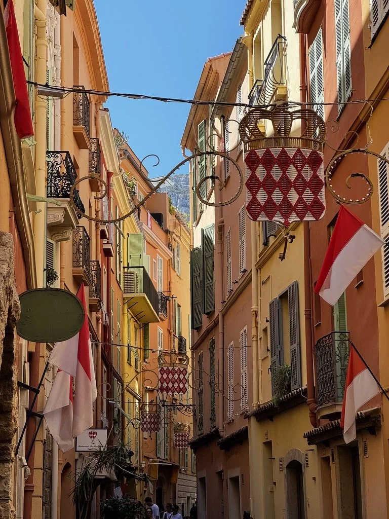 Narrow European street lined with pastel-colored buildings, decorative signs, and Croatian flags, under a bright blue sky.